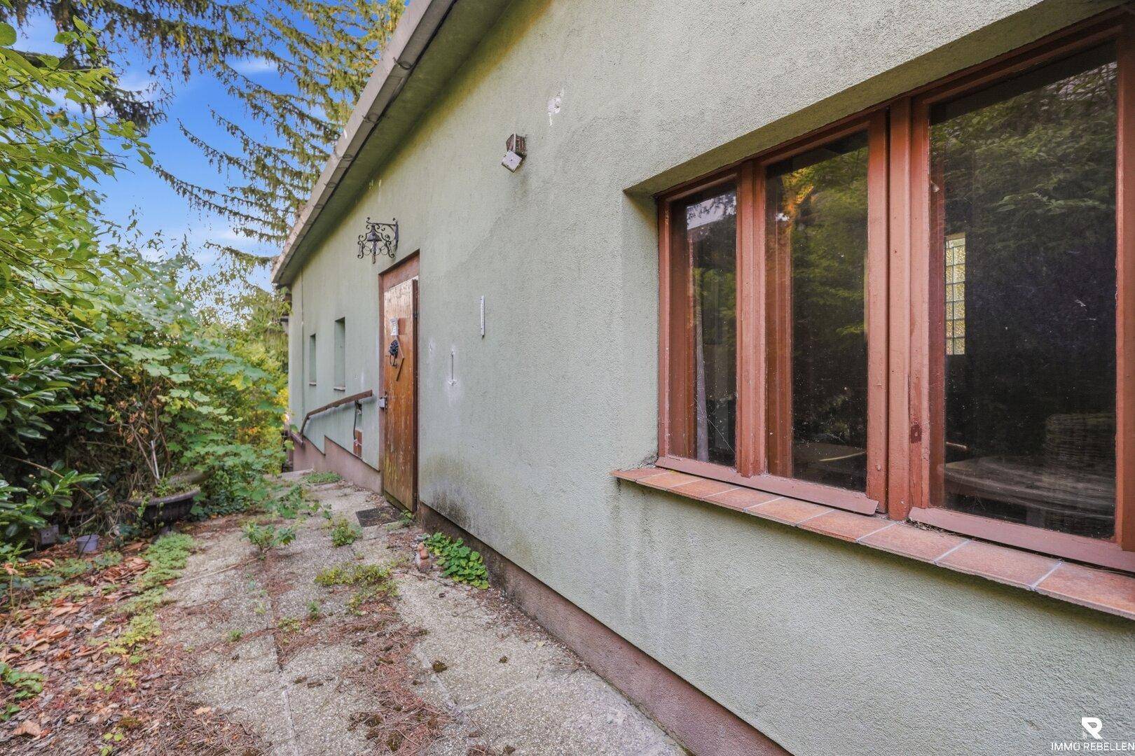Seitenansicht des Hauses mit grüner Fassade, Holzfenstern und einem gepflasterten Weg.