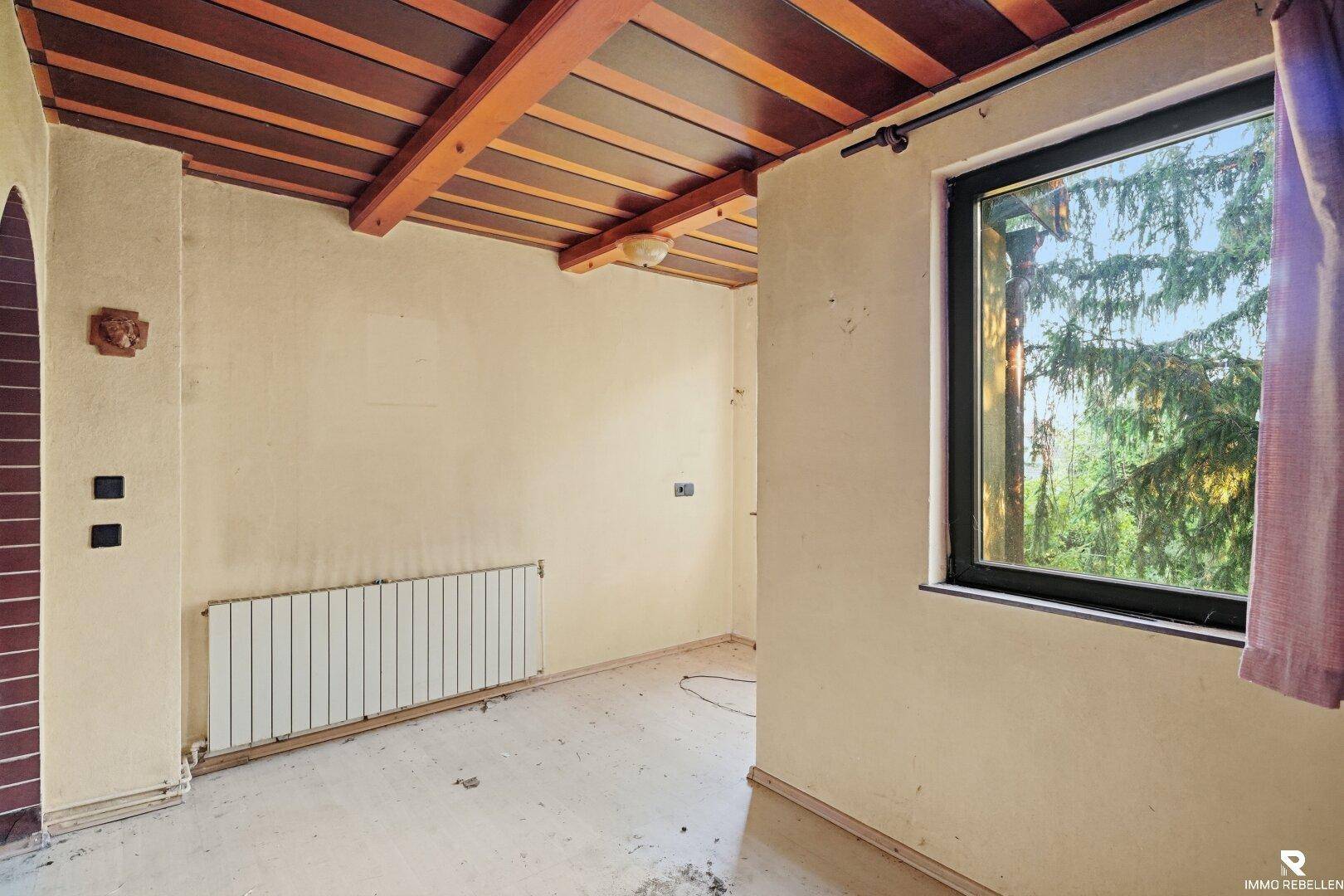 Leerer Raum mit Holzbalkendecke, Fenster und Heizkörper, bereit zur Renovierung.