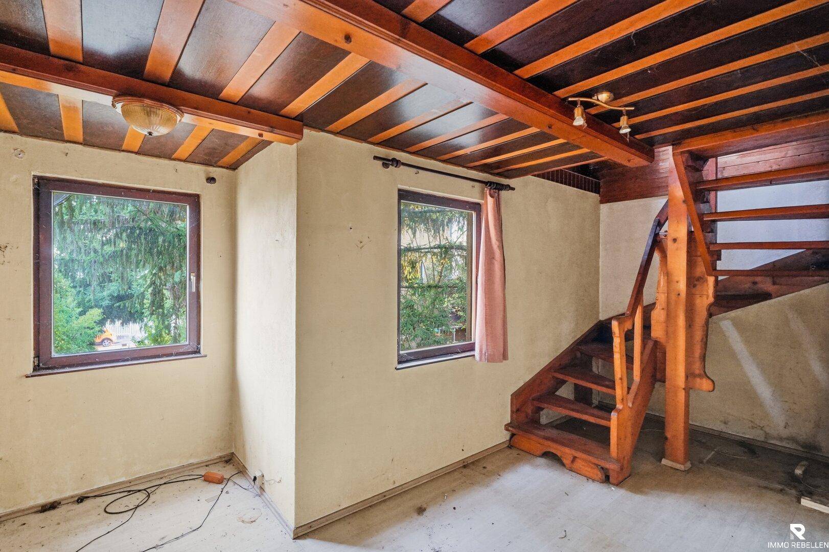 Raum im Originalzustand mit Holzdecke, zwei Fenstern und Holztreppe, benötigt Renovierung.