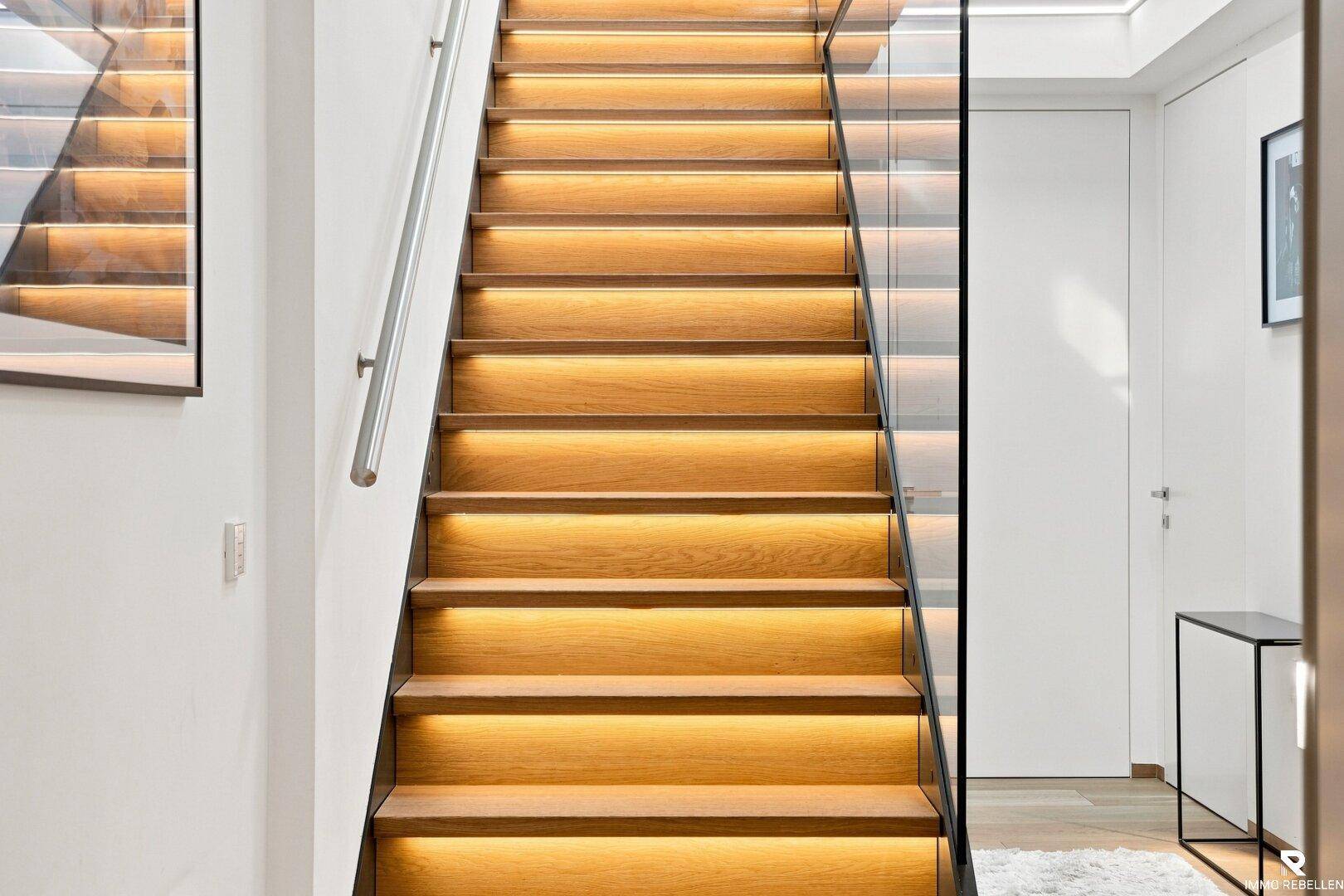 Beleuchtete Holztreppe mit Glasgeländer und modernem Design im Eingangsbereich.