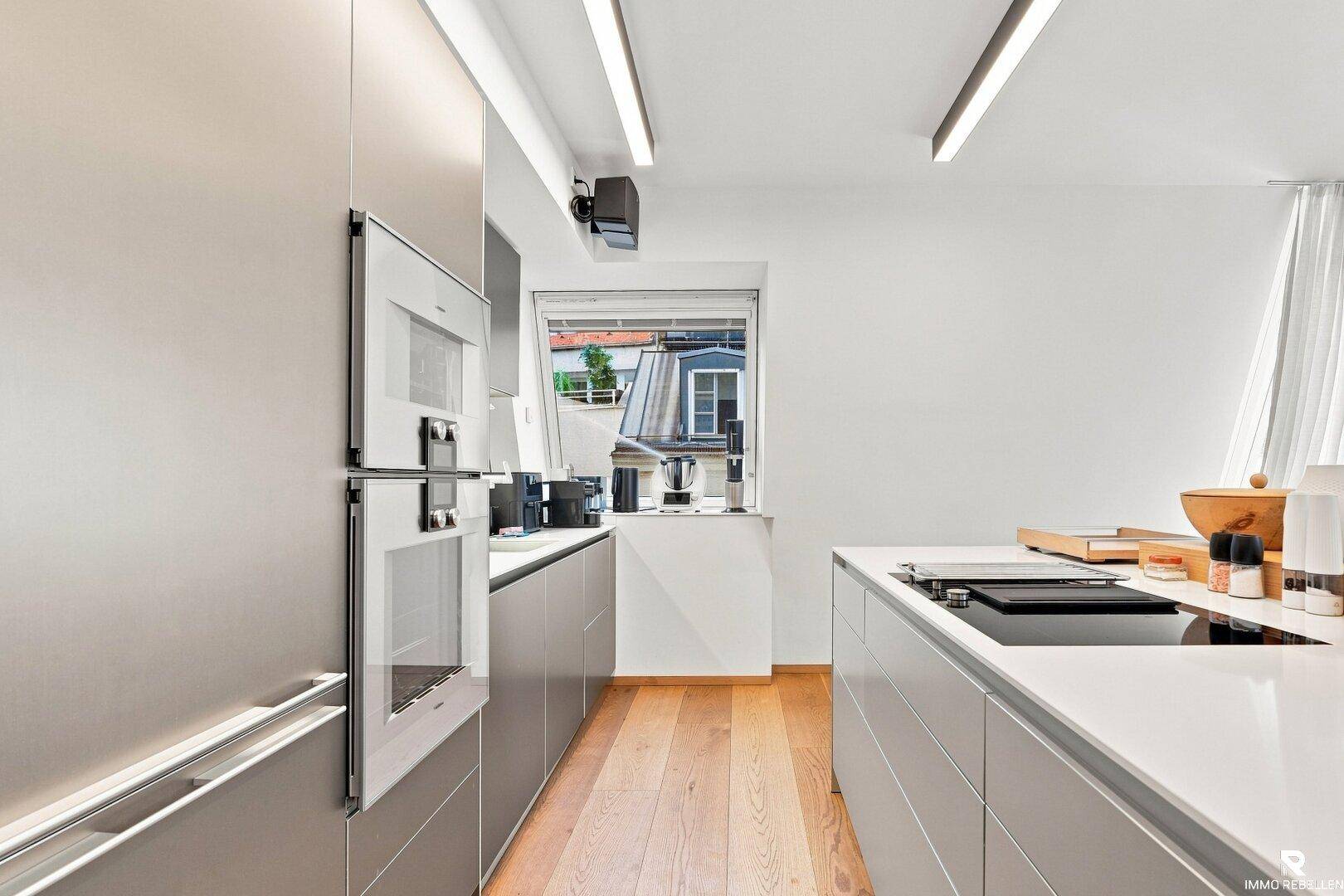 Moderne Küche mit grauen Schränken, weißen Arbeitsplatten und Blick nach draußen.