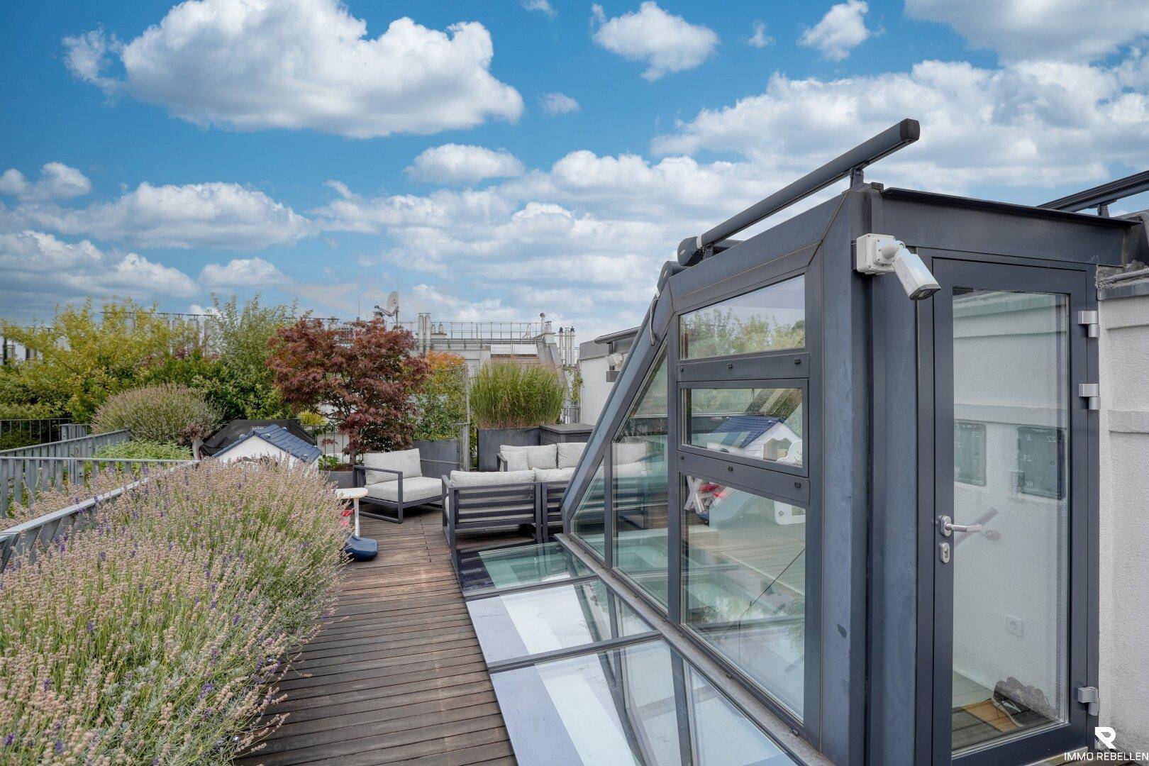 Geräumige Dachterrasse mit Holzboden, Glasaufbau und üppiger Bepflanzung unter blauem Himmel.