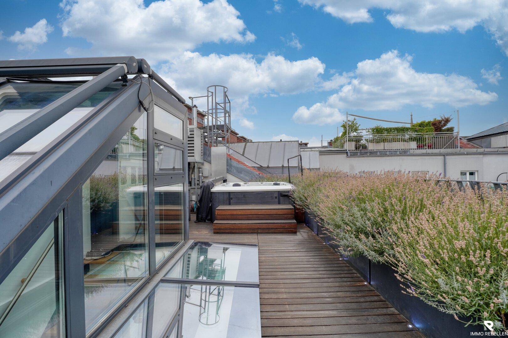 Dachterrasse mit Whirlpool, Holzboden und üppiger Bepflanzung unter blauem Himmel.