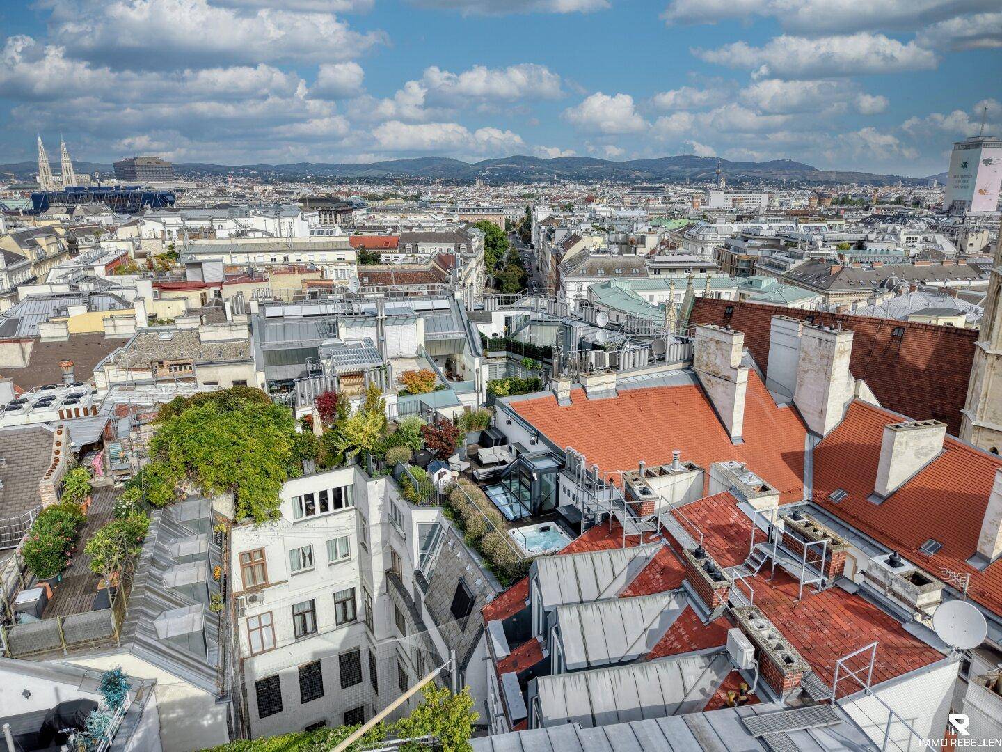 Panoramablick über die Dächer der Stadt mit grünen Terrassen und einer Bergkulisse am Horizont.