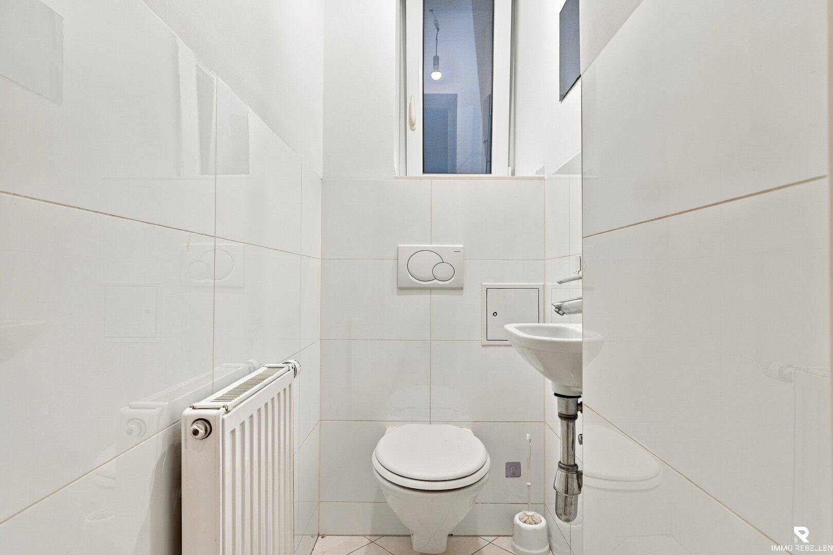Helles WC mit weißen Fliesen, wandhängender Toilette und kleinem Waschbecken.