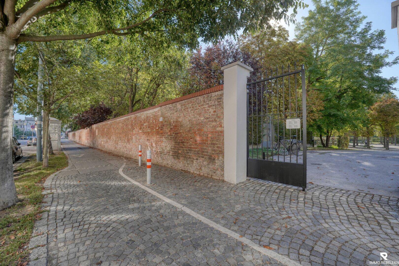 Historische Ziegelmauer mit einem schmiedeeisernen Tor, grenzt an einen gepflasterten Gehweg.