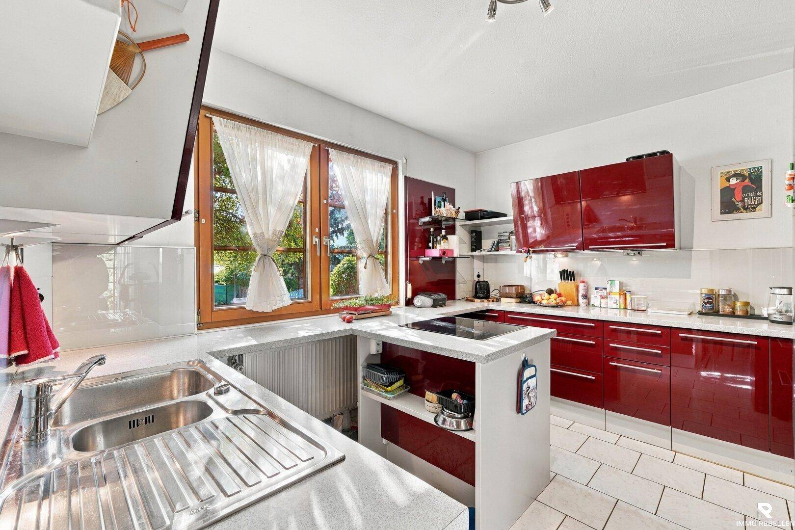 Moderne Küche mit glänzenden roten Fronten, Kochinsel und Fenster mit Blick ins Grüne.