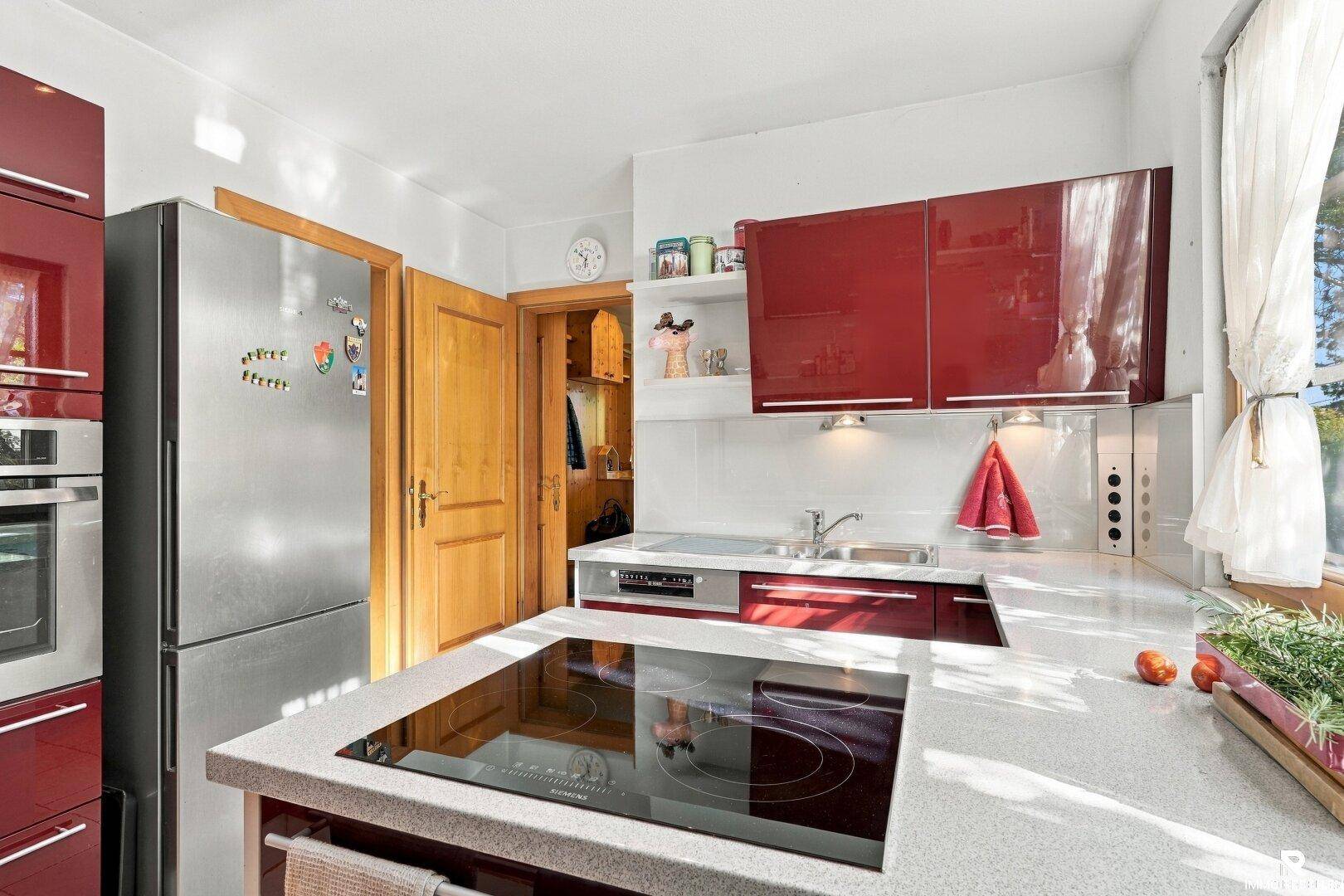 Funktionale Küche mit roten Schränken, Kühlschrank und Zugang zu einem weiteren Raum.
