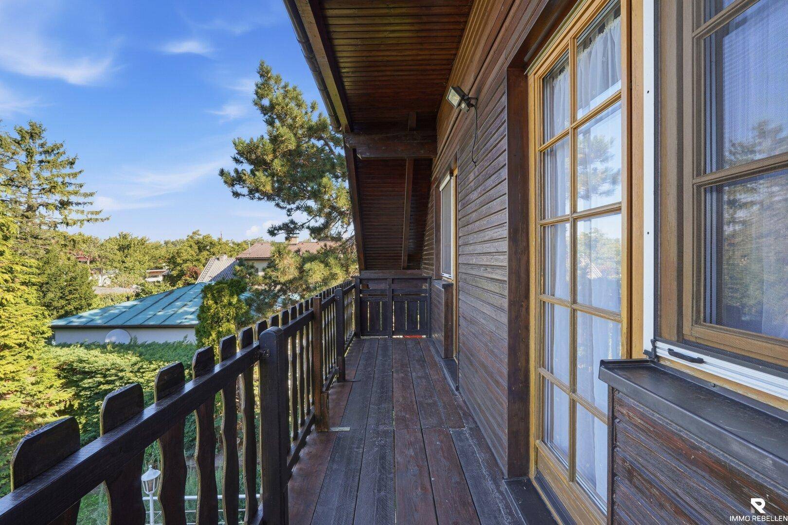 Langer Holzbalkon mit Blick auf Bäume und Nachbarhäuser unter blauem Himmel.