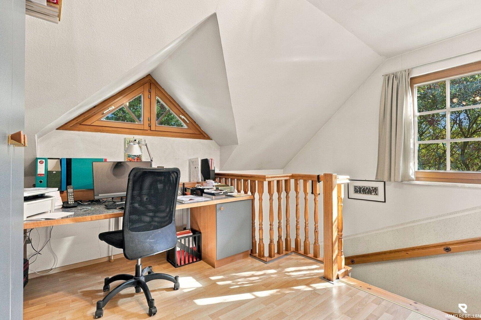 Arbeitsbereich im Dachgeschoss mit Schreibtisch, Holzboden und dreieckigem Fenster.