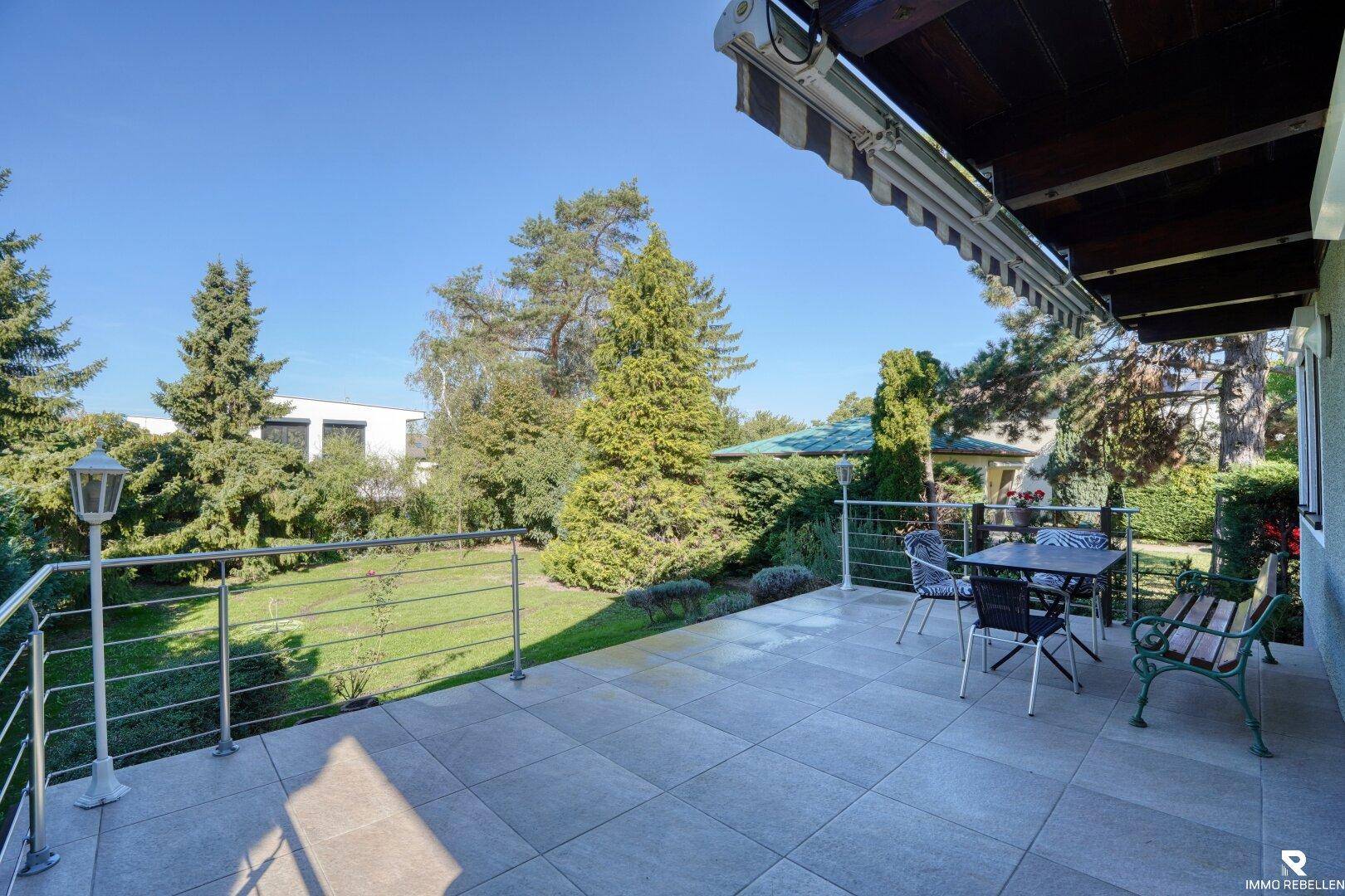 Große geflieste Terrasse mit Markise, Gartenmöbeln und Blick auf den grünen Garten.