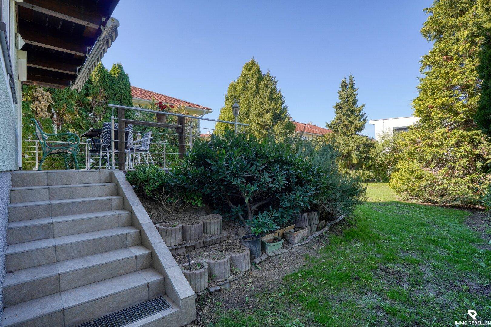 Gartenbereich mit Steintreppe zur Terrasse und üppiger Bepflanzung unter blauem Himmel.