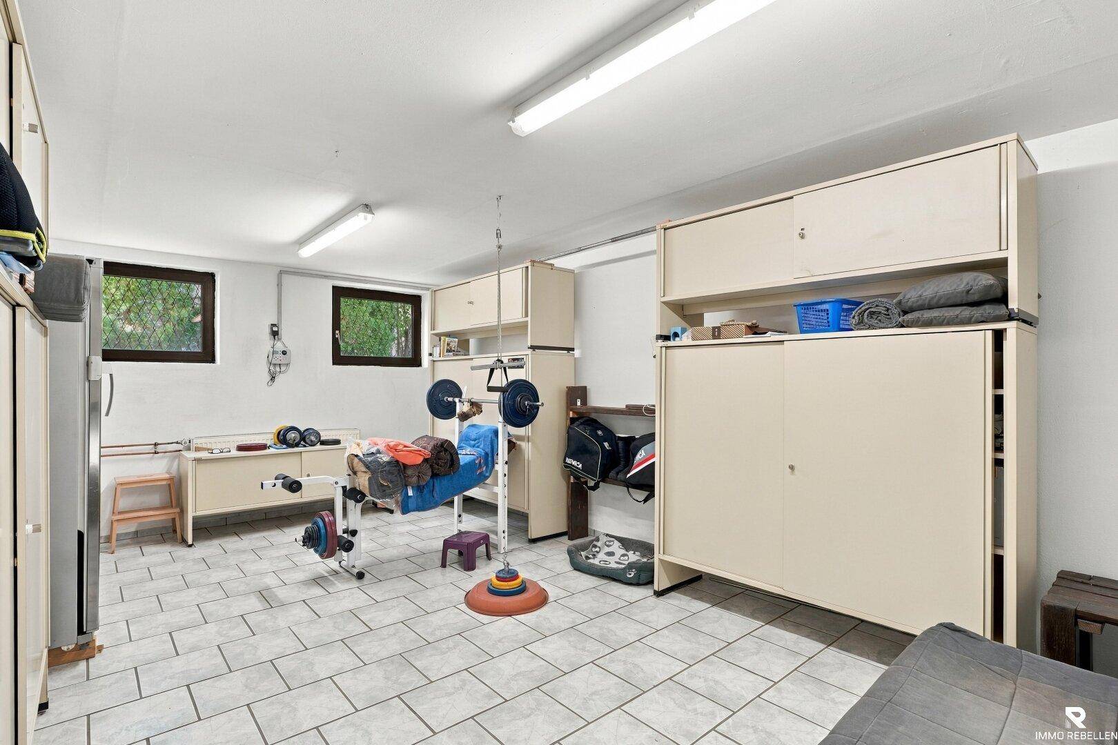 Fitnessbereich im Keller mit Trainingsgeräten, Schränken und zwei kleinen Fenstern für Belüftung.