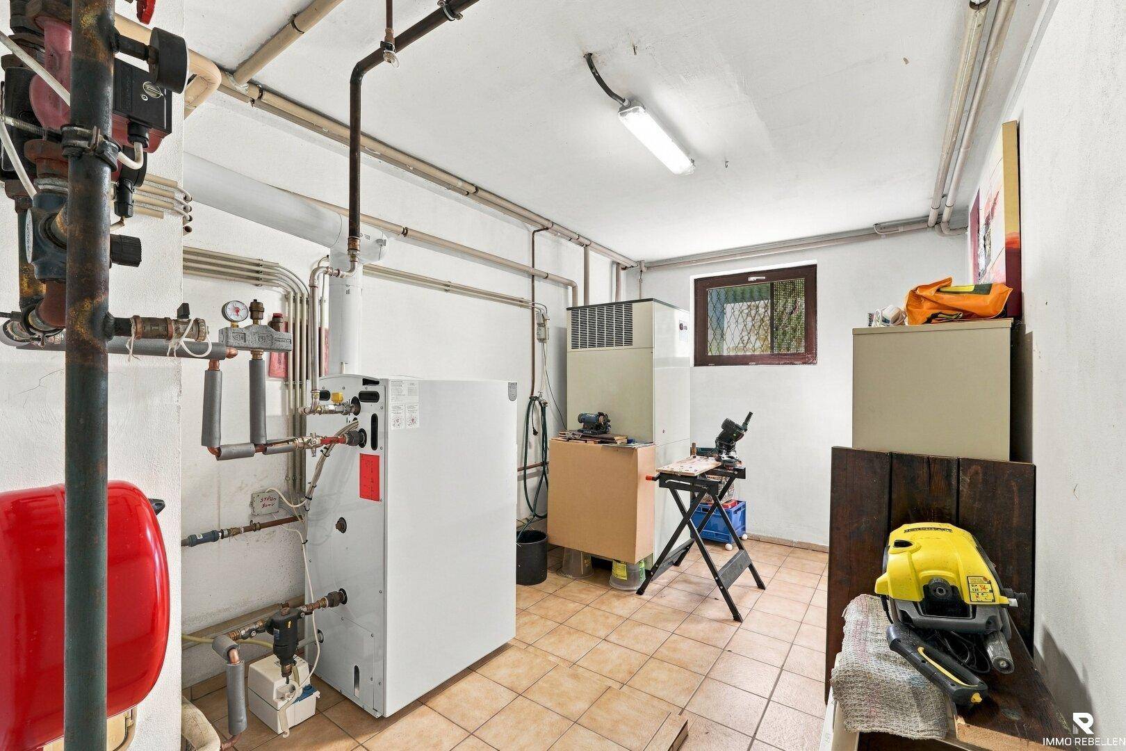 Heizraum im Keller mit Heizungsanlage, Rohren und einem kleinen Fenster für natürliche Belüftung.