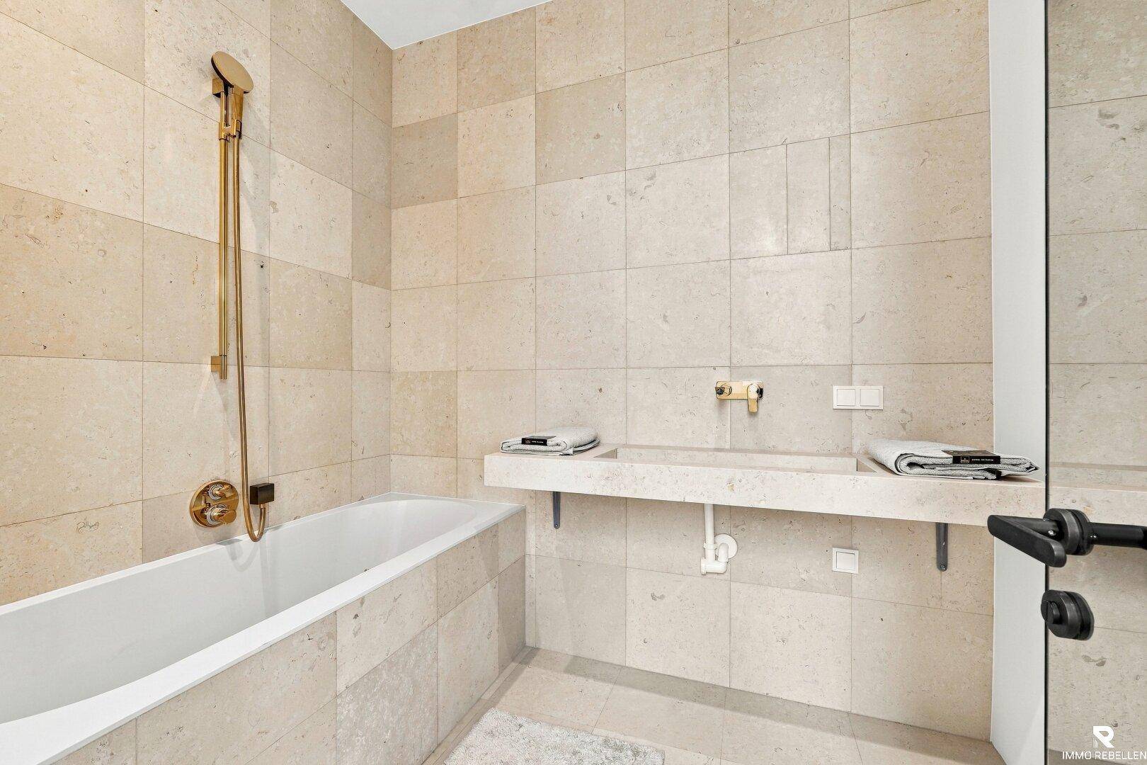 Modernes Badezimmer mit beigefarbenen Fliesen, Badewanne und goldfarbenen Armaturen.