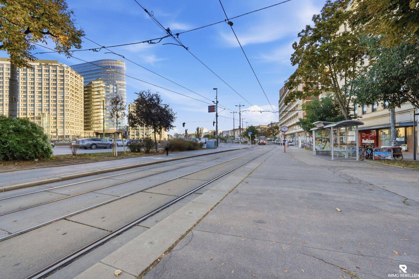 Breite Straße mit Straßenbahnschienen und modernen Gebäuden im Hintergrund.