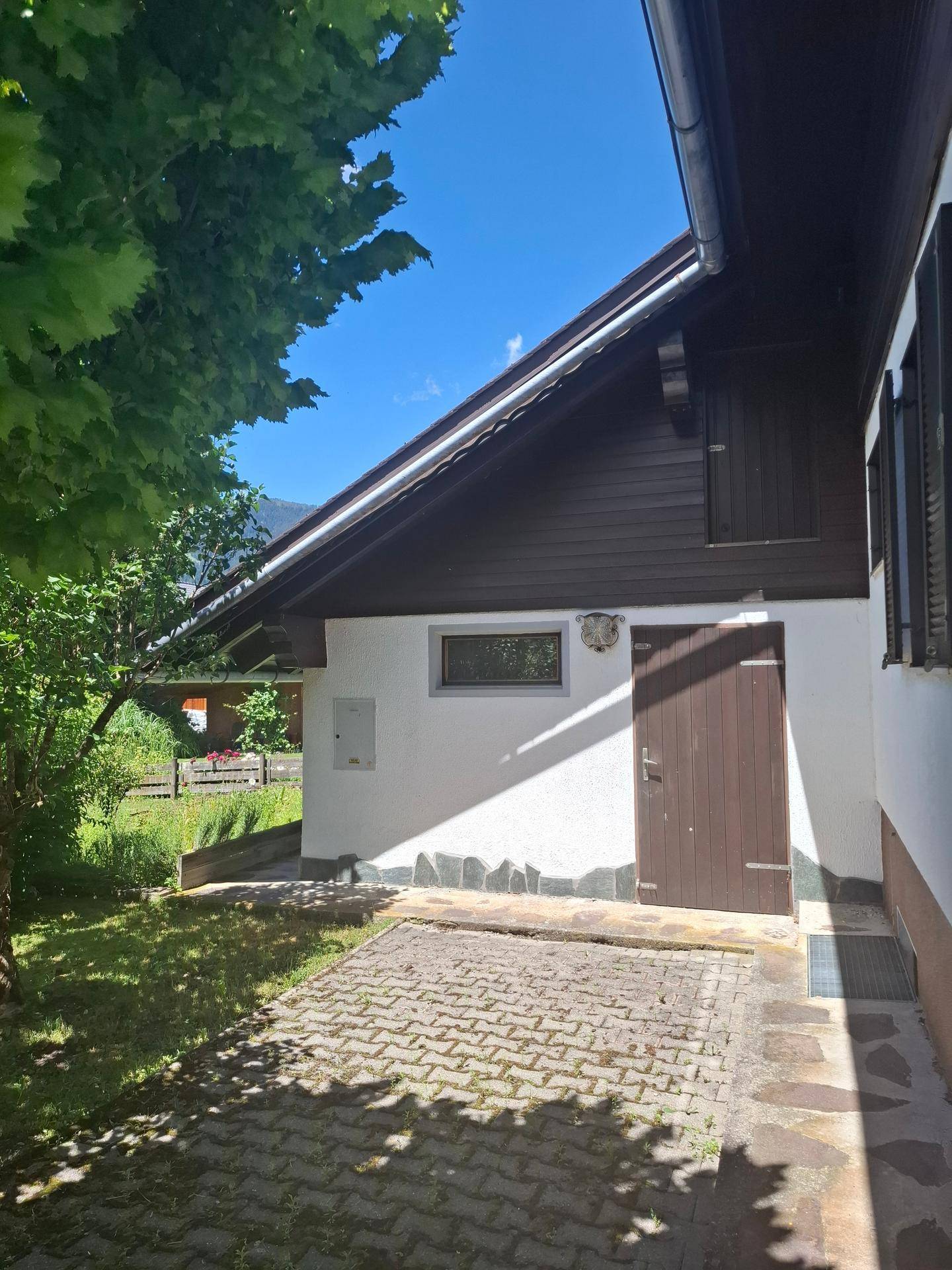 Seitenansicht des Hauses mit einer Tür und einem kleinen Fenster, daneben ein gepflasterter Bereich.