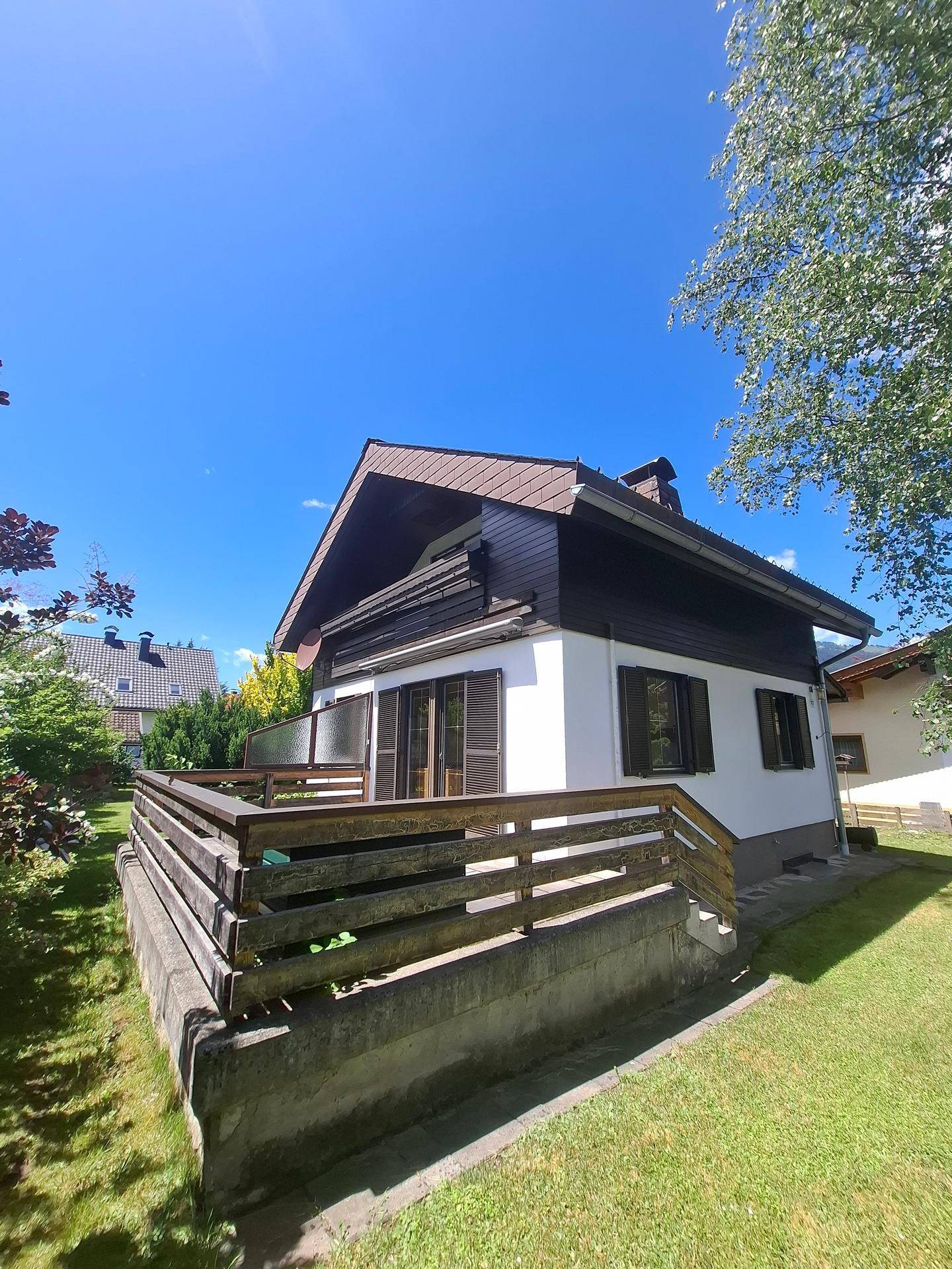 Seitliche Ansicht des Hauses mit einer erhöhten Terrasse und Holzgeländer, umgeben von grünem Rasen.