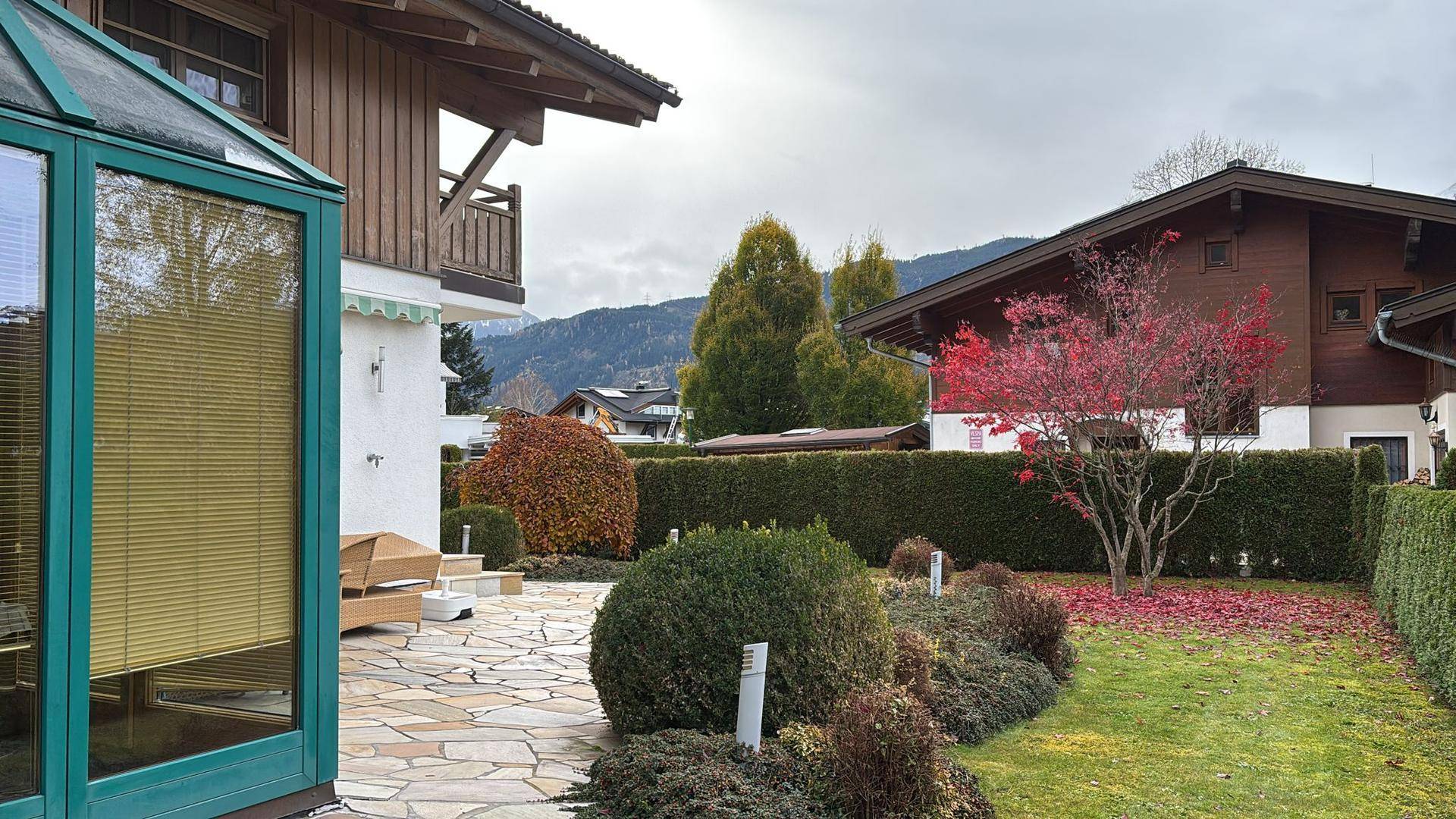 Gepflegter Gartenbereich mit Terrasse, Zierpflanzen und einem Wintergarten am Haus mit Bergblick.