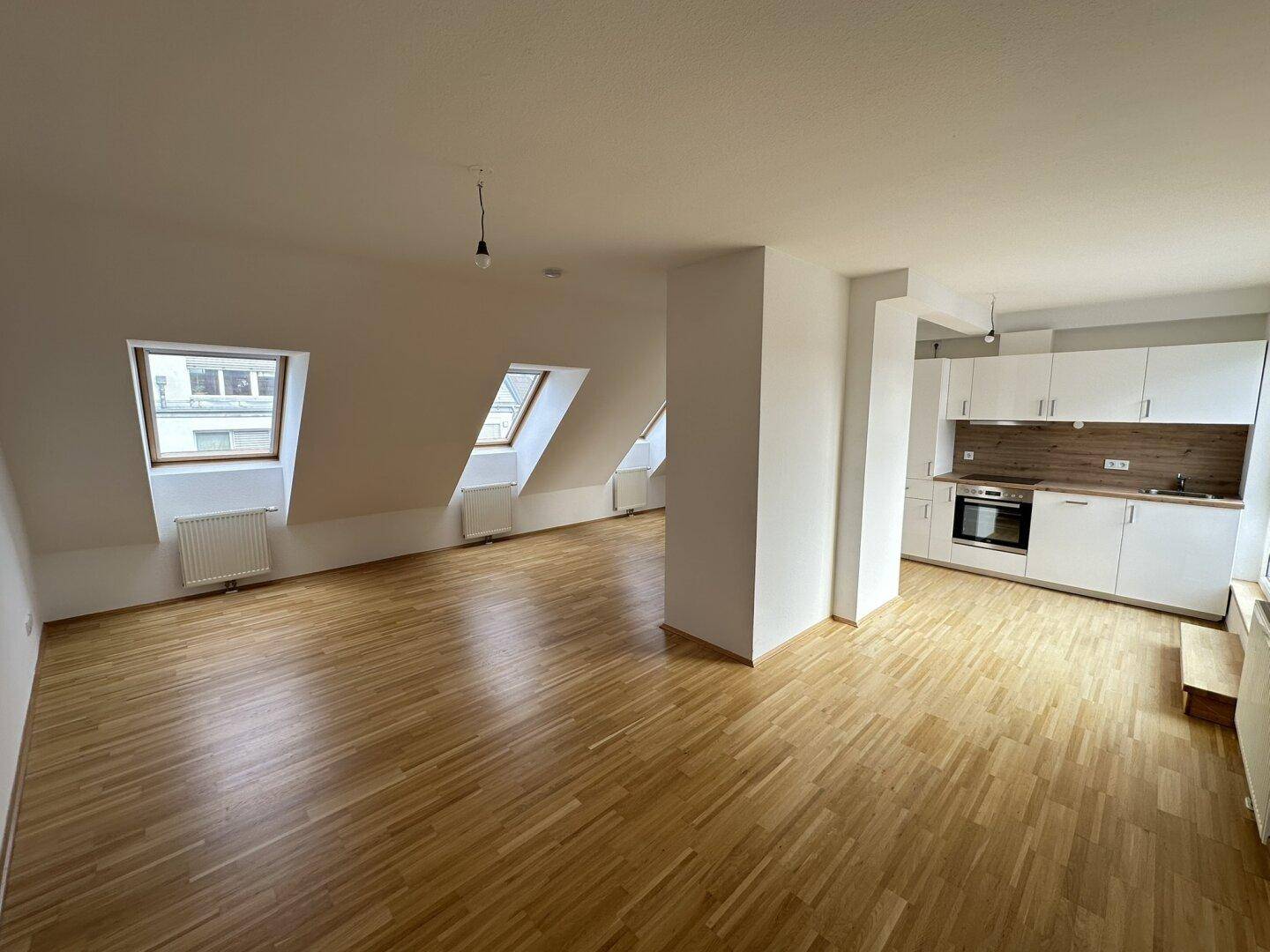 Geräumiger Wohnbereich mit Holzboden, Dachschrägen und offener Küche im Hintergrund.