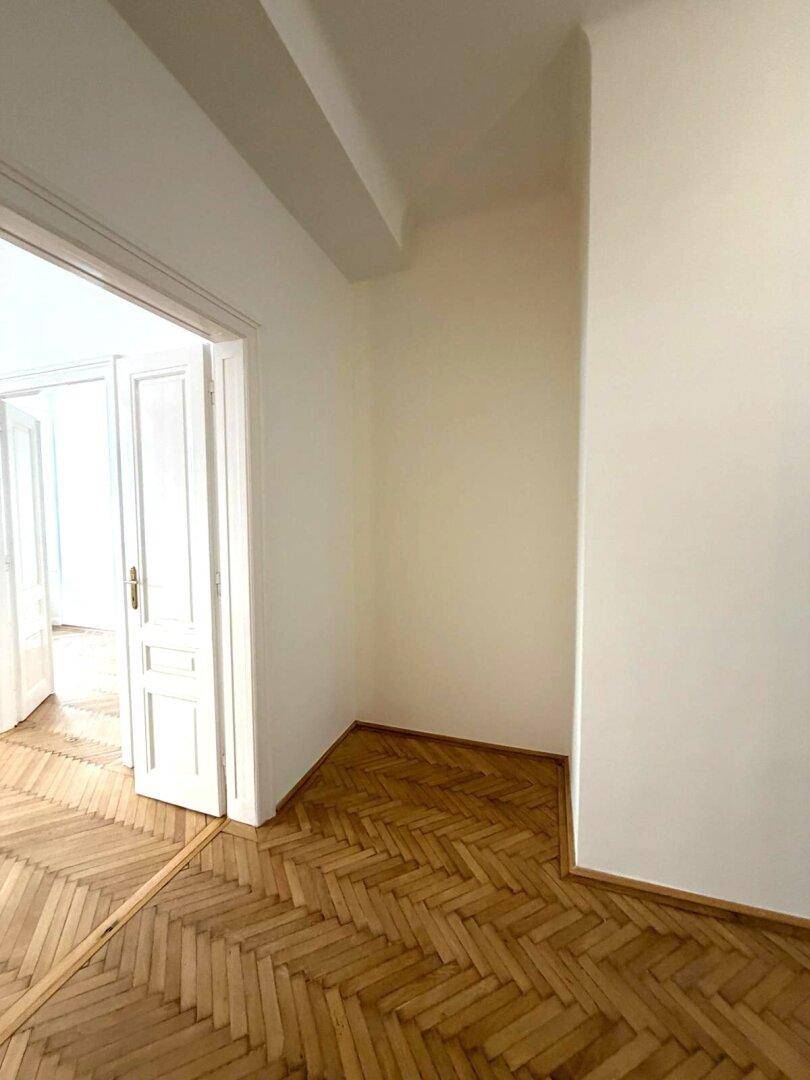 Ecke eines hellen Zimmers mit Fischgrätparkett und weiß gestrichenen Wänden.