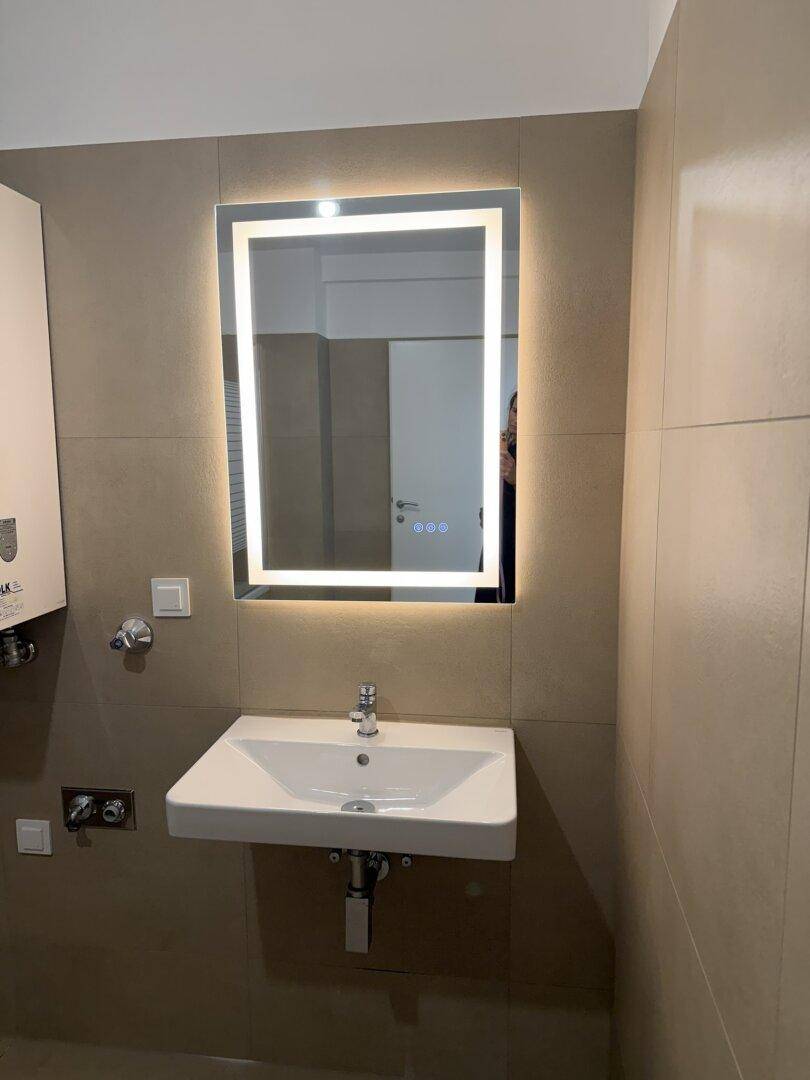 Badezimmer mit modernem Waschbecken, beleuchtetem Spiegel und beigefarbenen Wandfliesen.