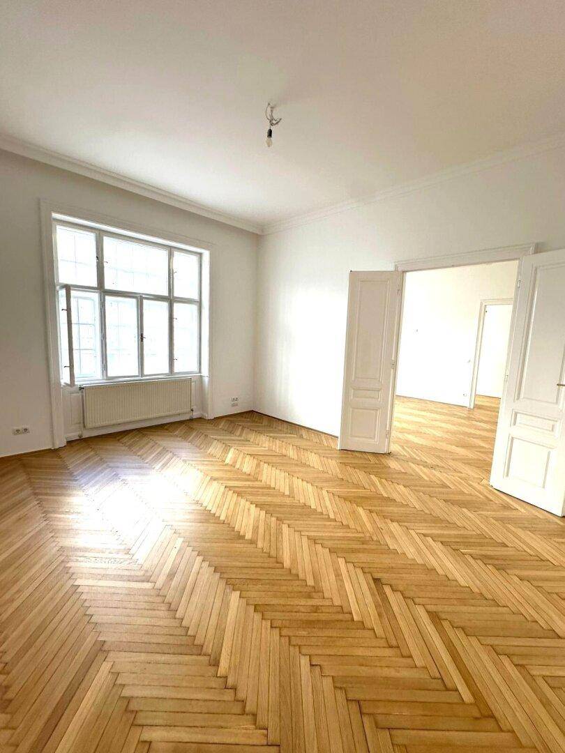 Lichtdurchflutetes Zimmer mit klassischem Fischgrätparkett und hohen Decken, ideal für Wohnzwecke.