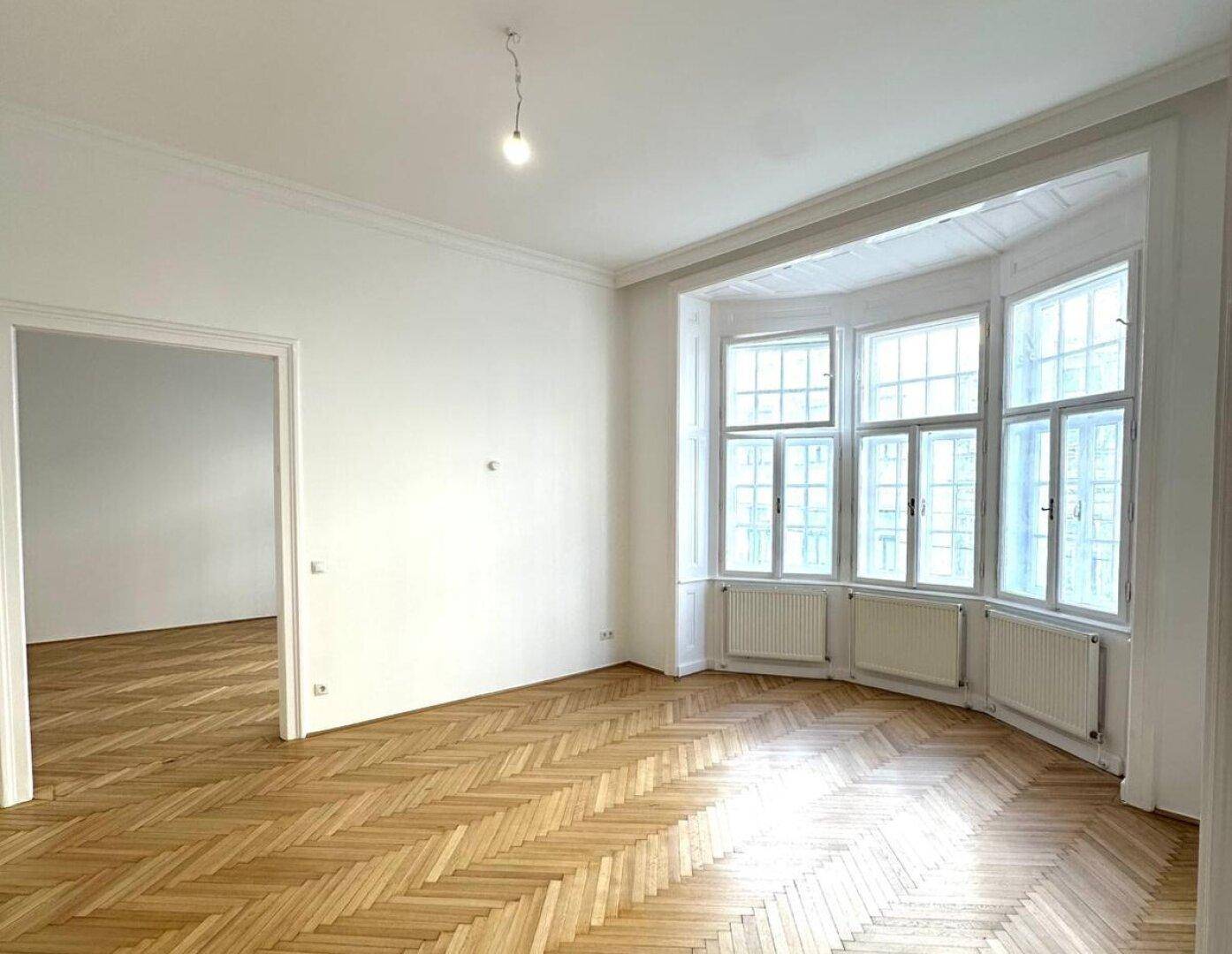Geräumiges Zimmer mit Fischgrätparkett und einem Erkerfenster, das viel Licht hereinlässt.