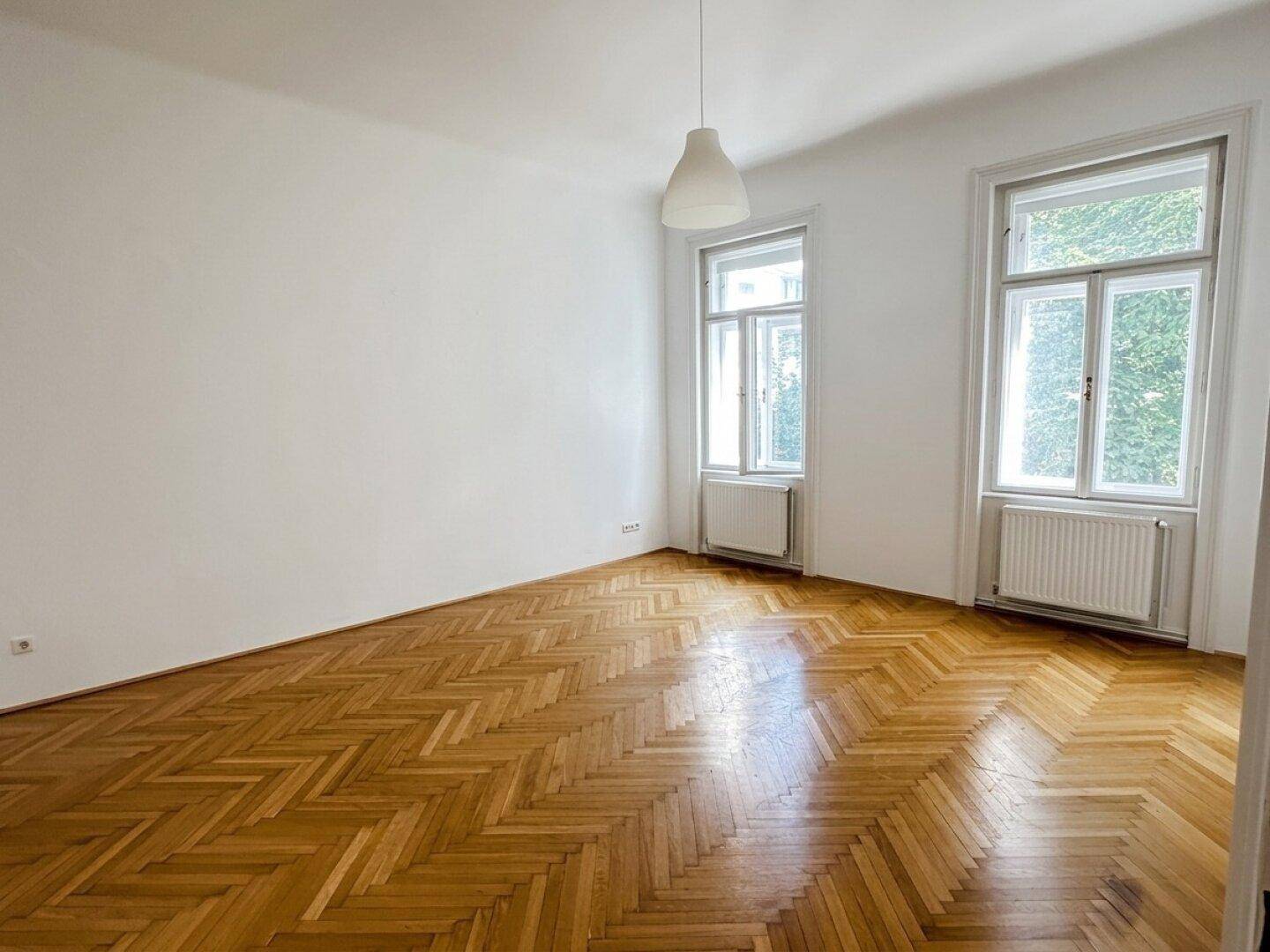 Helles Zimmer mit Fischgrätparkett und zwei großen Fenstern, die einen Blick ins Grüne bieten.