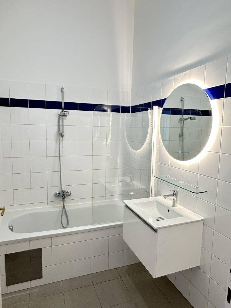 Modernes Badezimmer mit Badewanne, beleuchtetem Spiegel und weißen Fliesen mit blauer Akzentlinie.