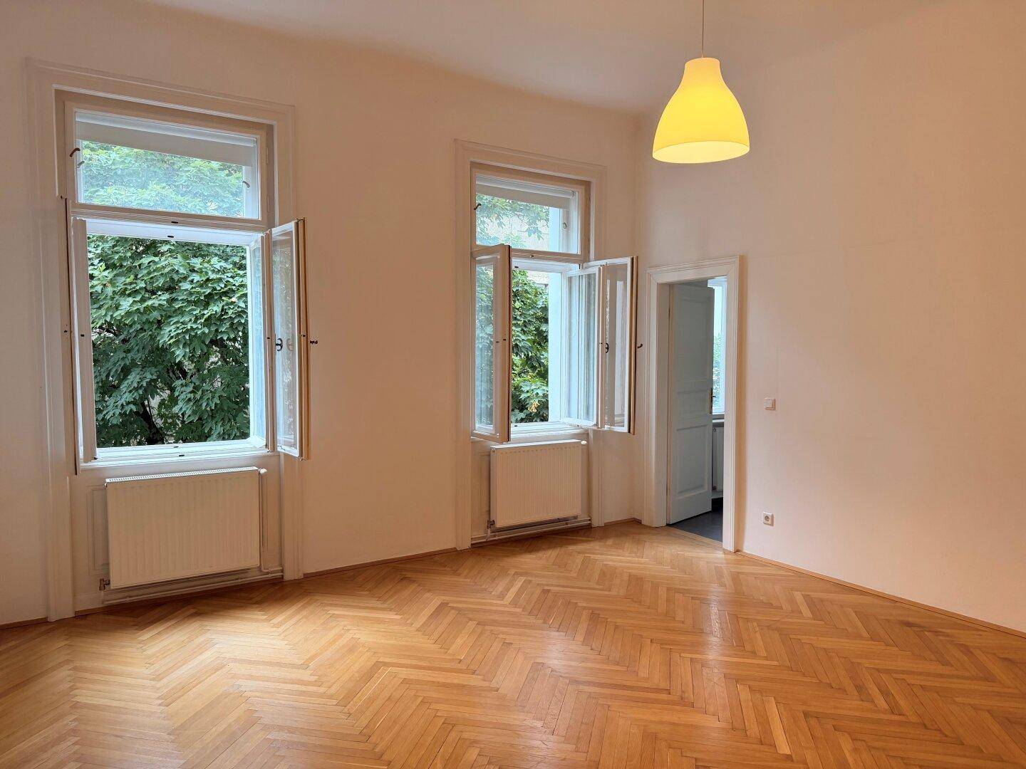 Helles Wohnzimmer mit elegantem Fischgrätparkett und Blick ins Grüne durch hohe Fenster.