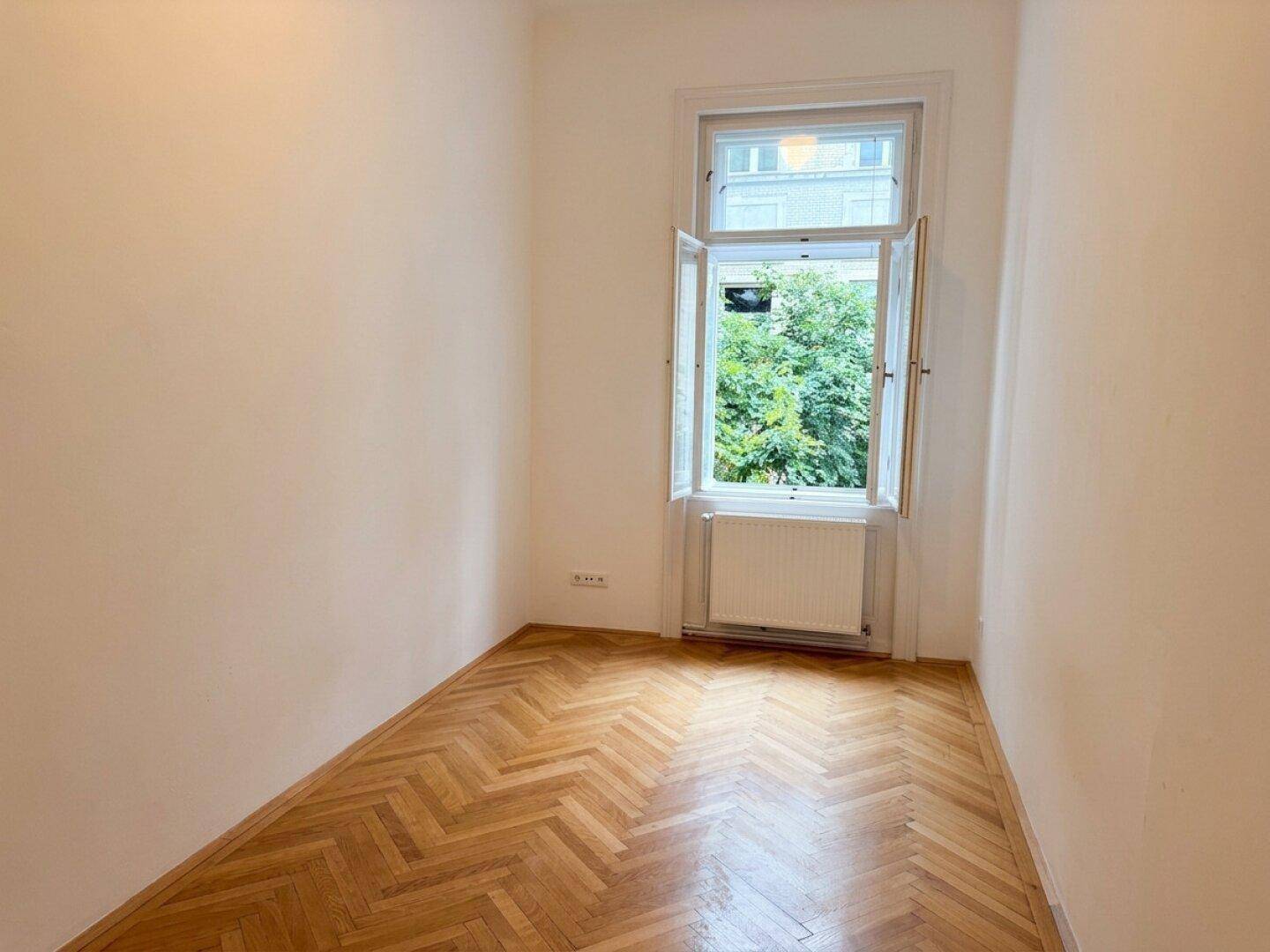 Kleineres Zimmer mit Fischgrätparkett und einem Fenster mit Blick auf Bäume.