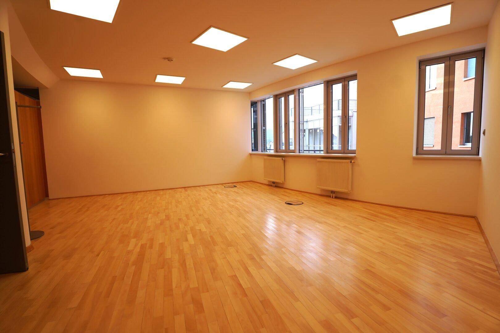 Leerer Büroraum mit Holzparkettboden und großen Fenstern, die viel Licht spenden.