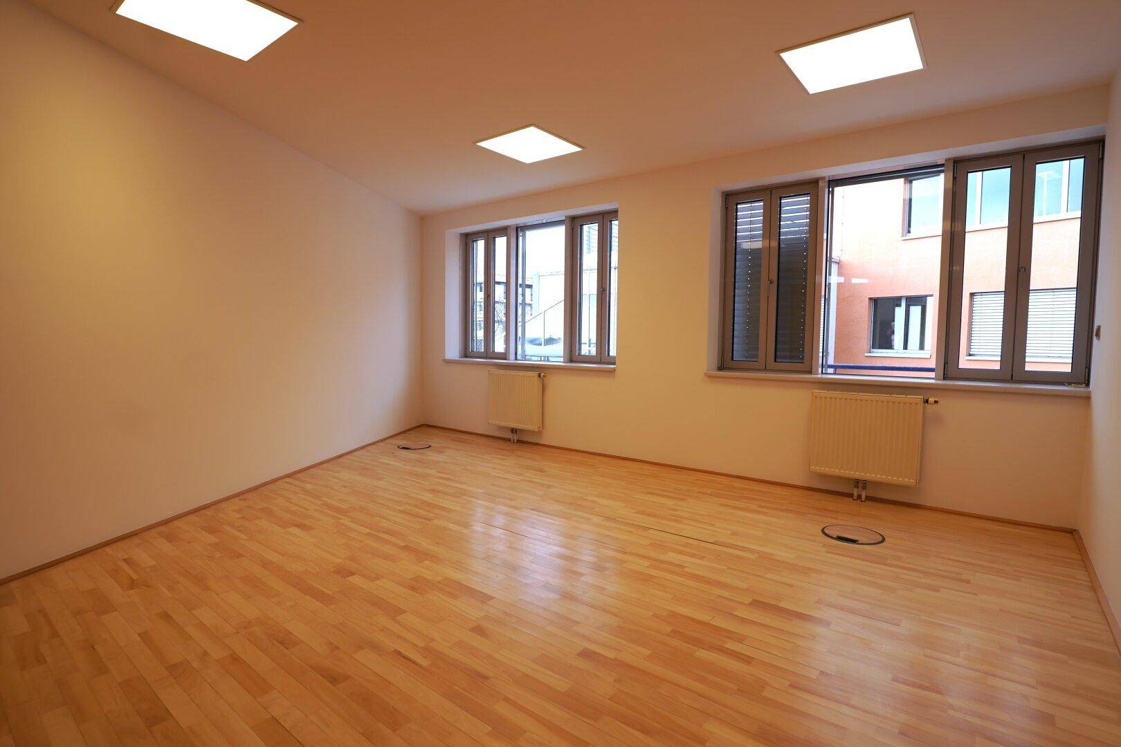 Leerer Büroraum mit Holzparkettboden und mehreren Fenstern, die Tageslicht hereinlassen.