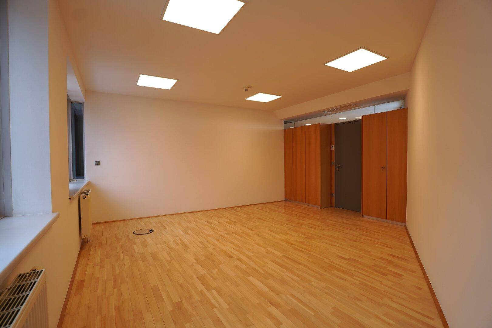 Leerer Büroraum mit diagonal verlegtem Holzparkett und integrierten Schränken.