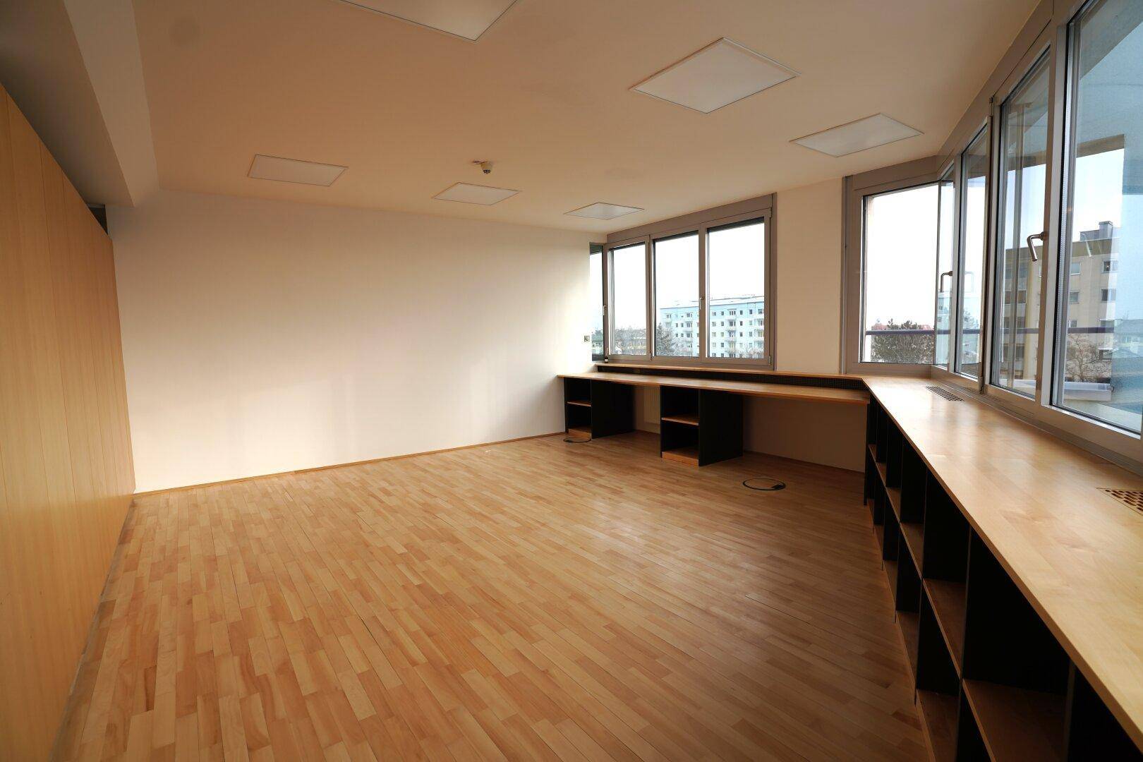 Leerer Büroraum mit großen Fenstern und integrierten Regalen entlang der Wand.