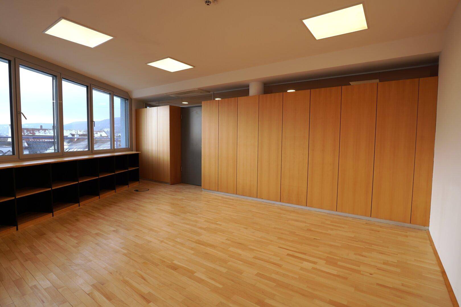 Leerer Büroraum mit großen Fenstern und integrierten Schränken entlang der Wand.