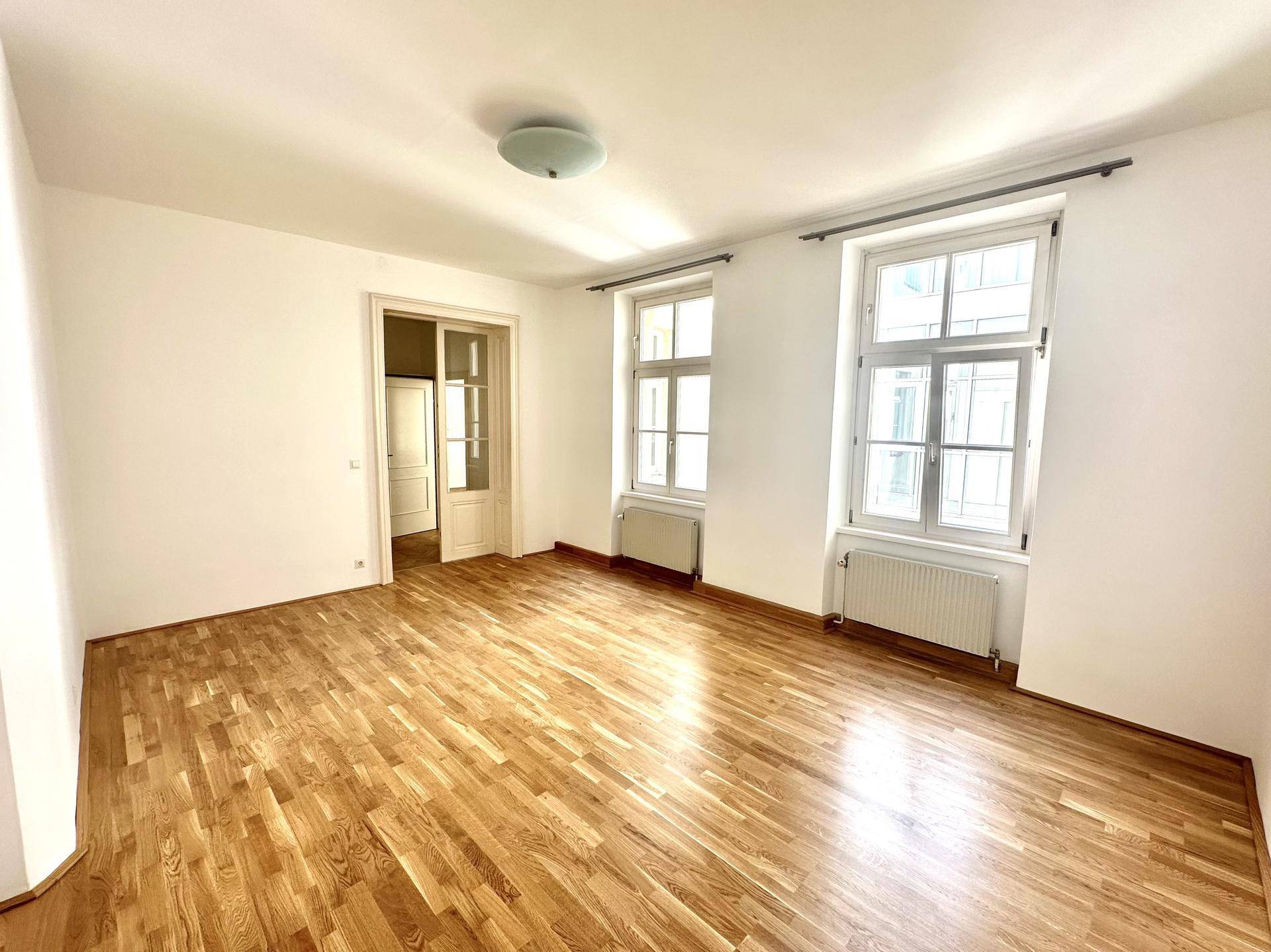 Lichtdurchflutetes Zimmer mit klassischem Fischgrätparkett und hohen Fenstern, bietet vielseitige Nutzungsmöglichkeiten.