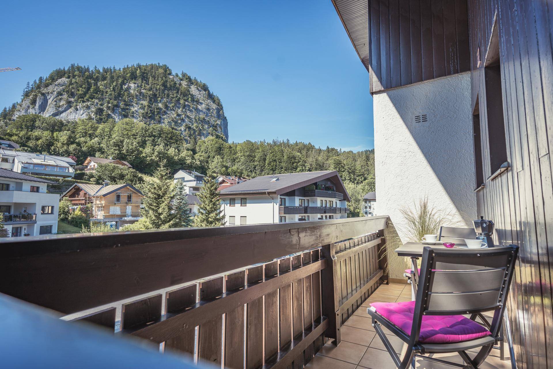 Gemütlicher Balkon mit kleinem Tisch und Stühlen, umgeben von grüner Natur und Bergen.