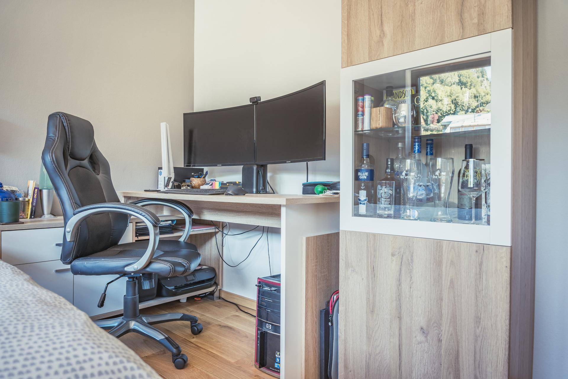 Funktionaler Arbeitsbereich mit zwei Monitoren, ergonomischem Stuhl und einem Schrank mit Glasvitrine.
