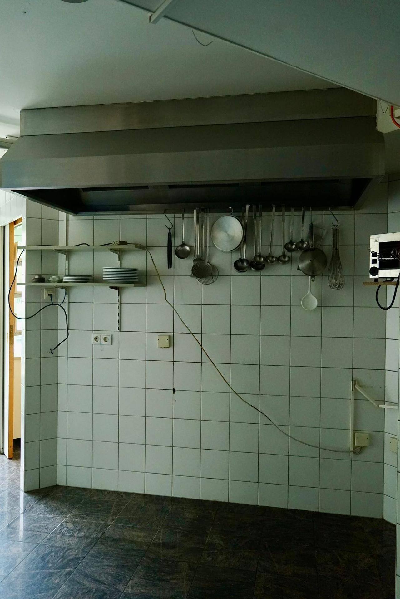 Gewerbliche Küche mit großer Dunstabzugshaube, gefliesten Wänden und aufgehängten Küchenutensilien.