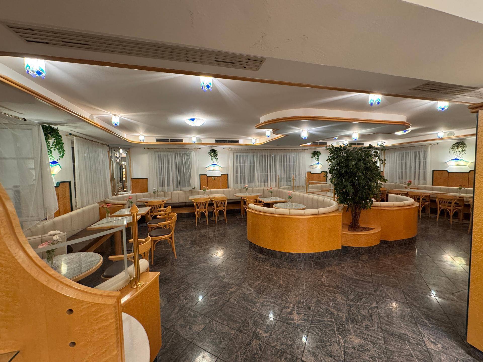 Geräumiger Café-Sitzbereich mit verschiedenen Tischen, Polsterbänken, dekorativer Beleuchtung und einer großen Pflanze.