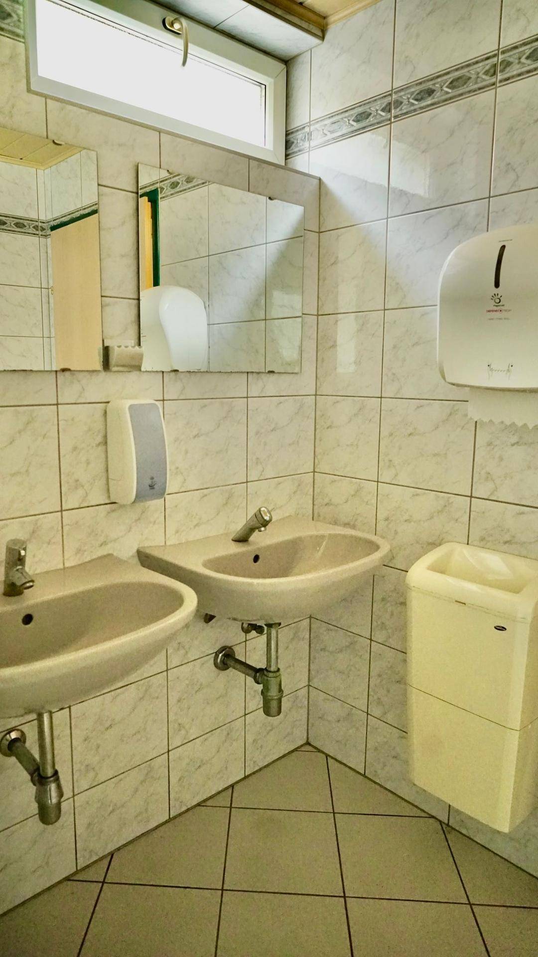 Öffentlicher Waschraum mit zwei Waschbecken, Spiegeln und weißen Fliesen an Wänden und Boden.