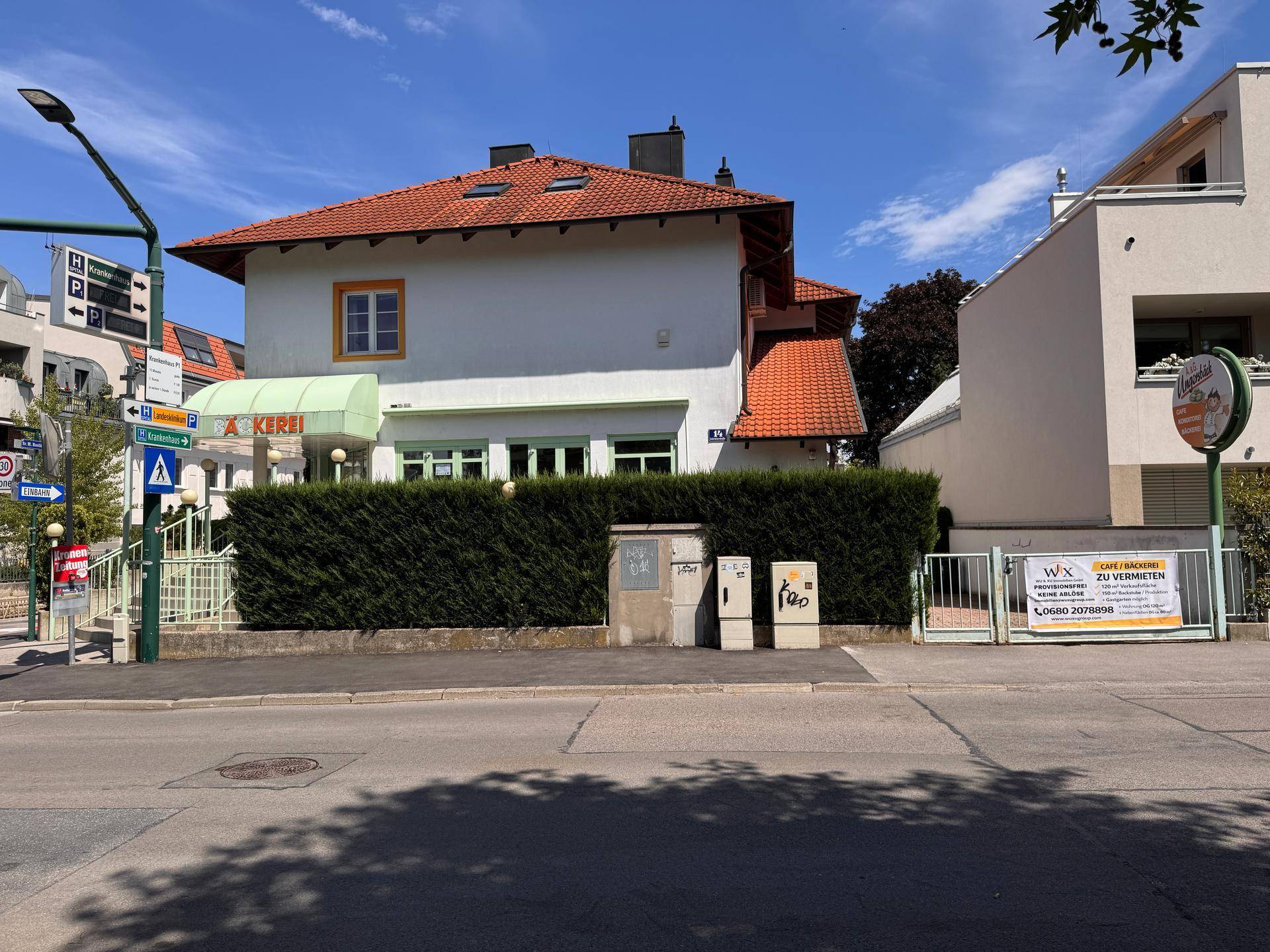 Außenansicht des Gebäudes mit rotem Ziegeldach und weißer Fassade, umgeben von einer Hecke und der Straße.