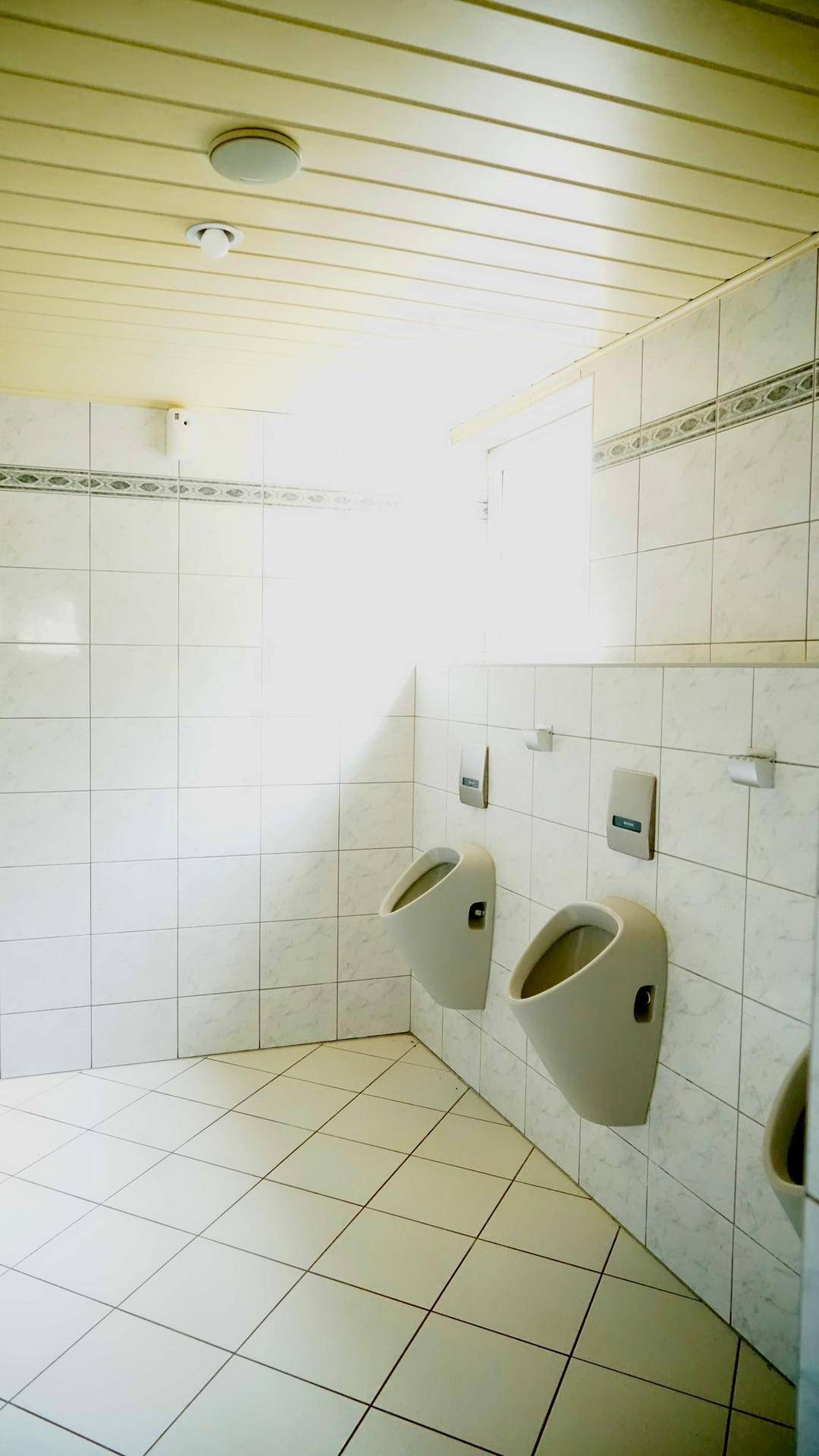 Herren-WC mit mehreren Urinalen, weißen Fliesen an Wänden und Boden sowie einem kleinen Fenster.