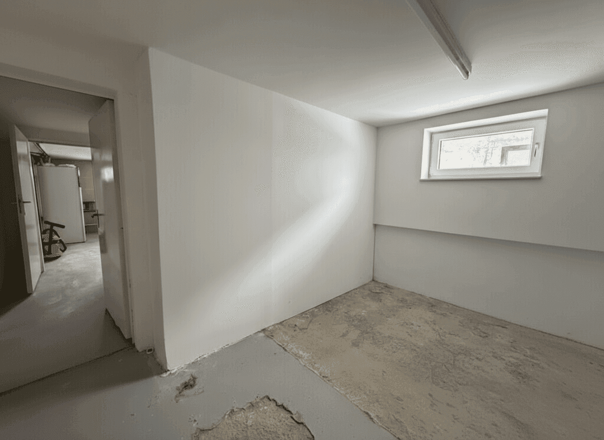 Kellerraum mit Betonboden, weiß gestrichenen Wänden und kleinem Fenster.