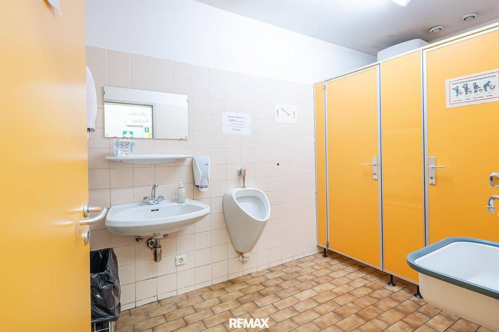 Öffentliche Toilette mit mehreren Kabinen, Urinal und Waschbecken, gefliestem Boden.
