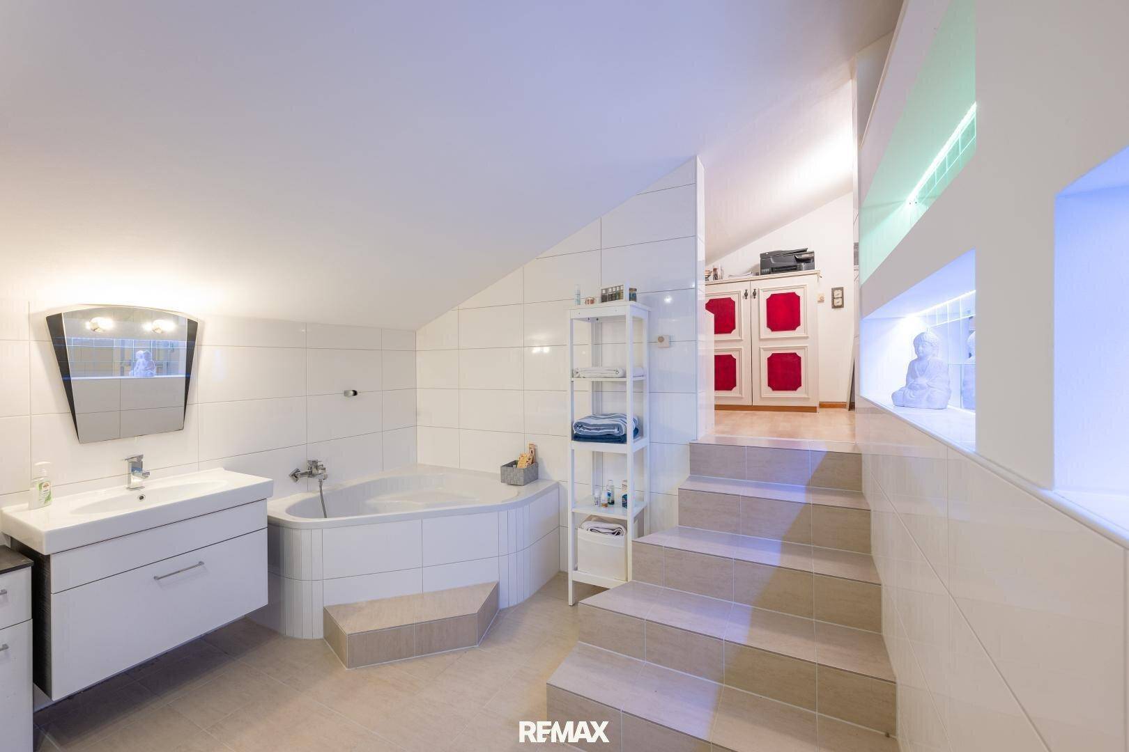 Helles Badezimmer mit Eckbadewanne, Waschtisch und Treppe zu einem erhöhten Bereich.