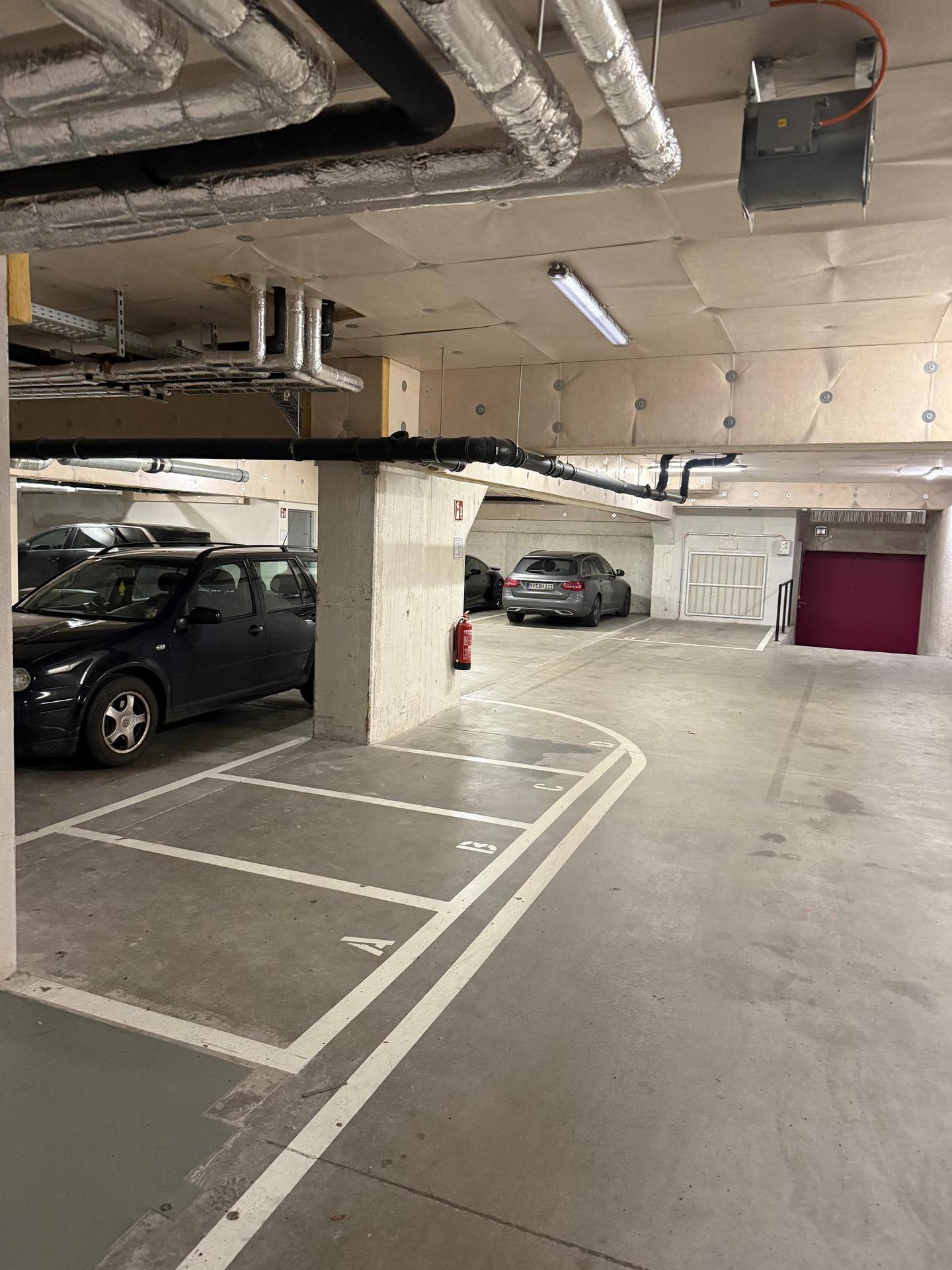 Tiefgarage mit markierten Stellplätzen, sichtbaren Rohren und geparkten Fahrzeugen.