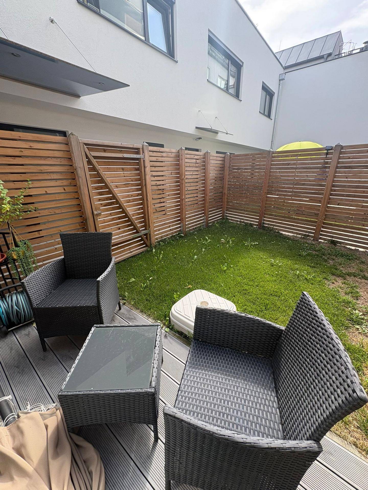 Außenbereich mit Holzterrasse, Sitzgelegenheiten und Rasenfläche, umgeben von einem Holzzaun.